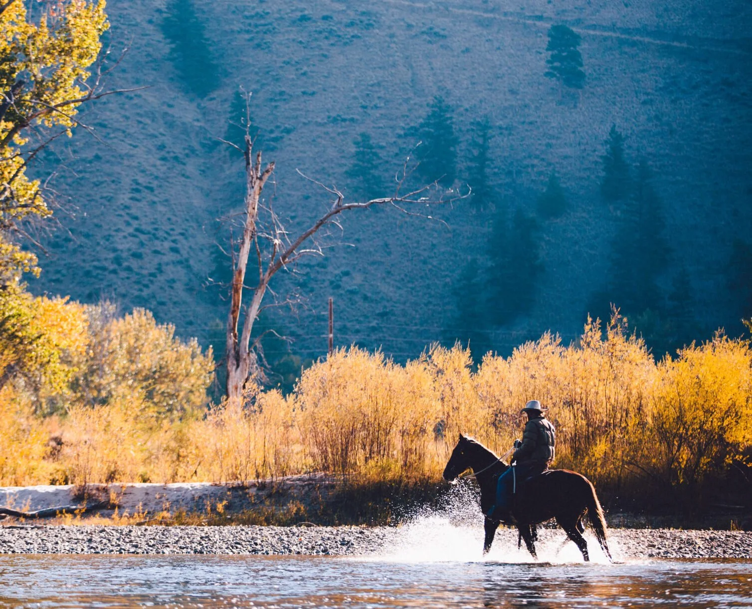 Person riding a horse through a shallow river with autumn foliage and mountains in the background.