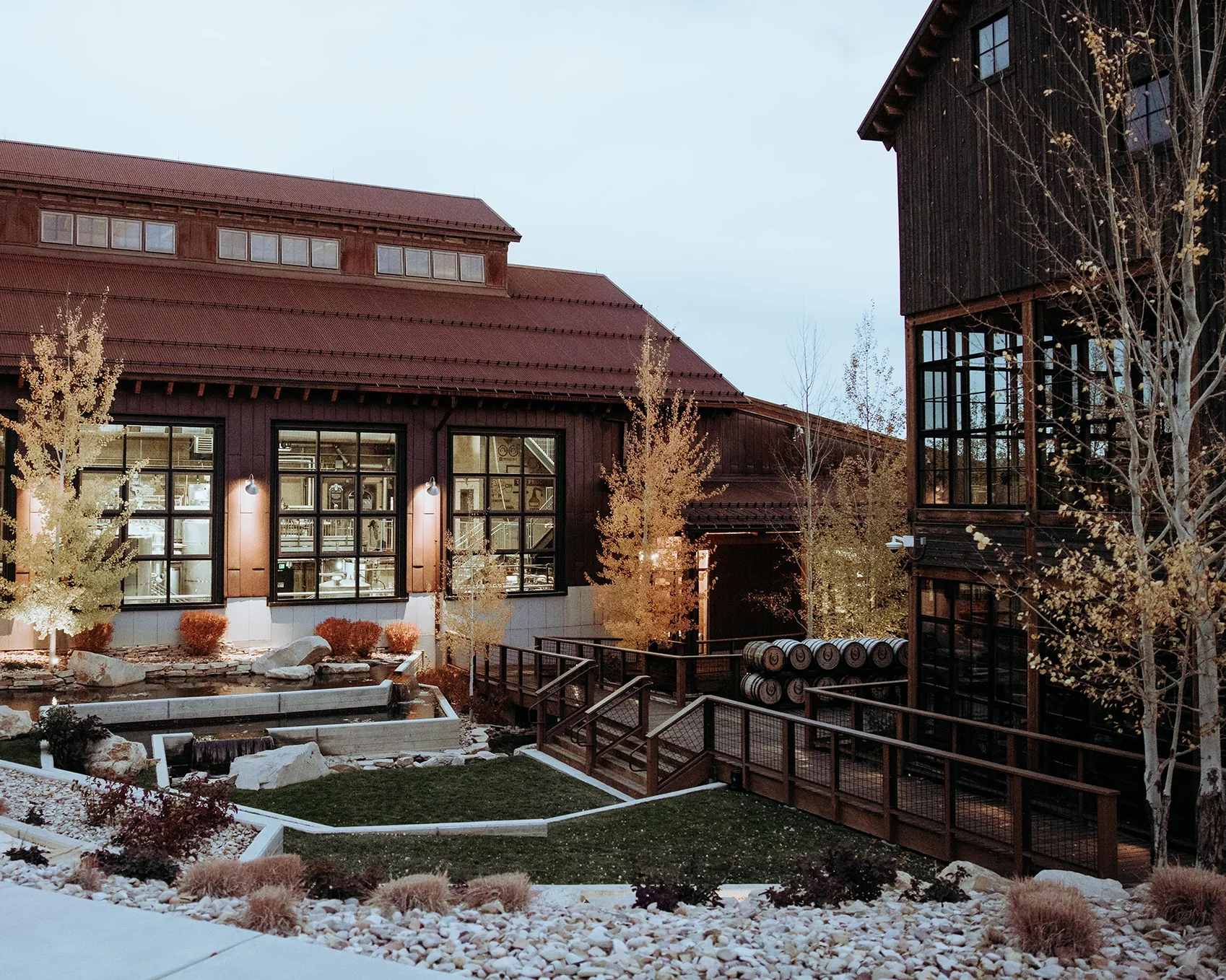 Exterior view of a modern building with large windows, surrounded by landscaped trees and rocks, with a wooden deck and barrels in the courtyard during dusk.