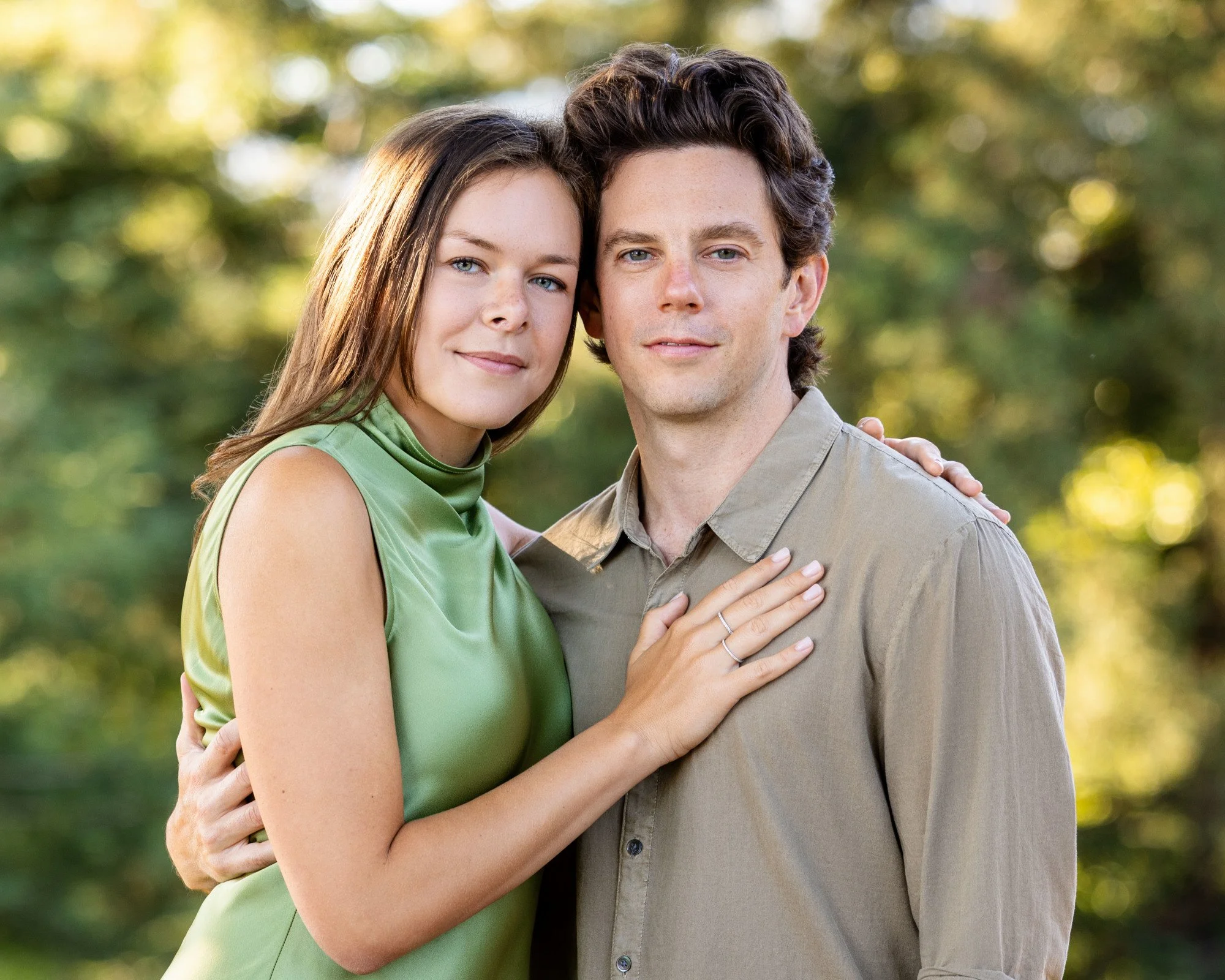 A young woman and young man standing close together outdoors, with the woman resting her hand on the man's chest, both looking at the camera, with blurred trees in the background.