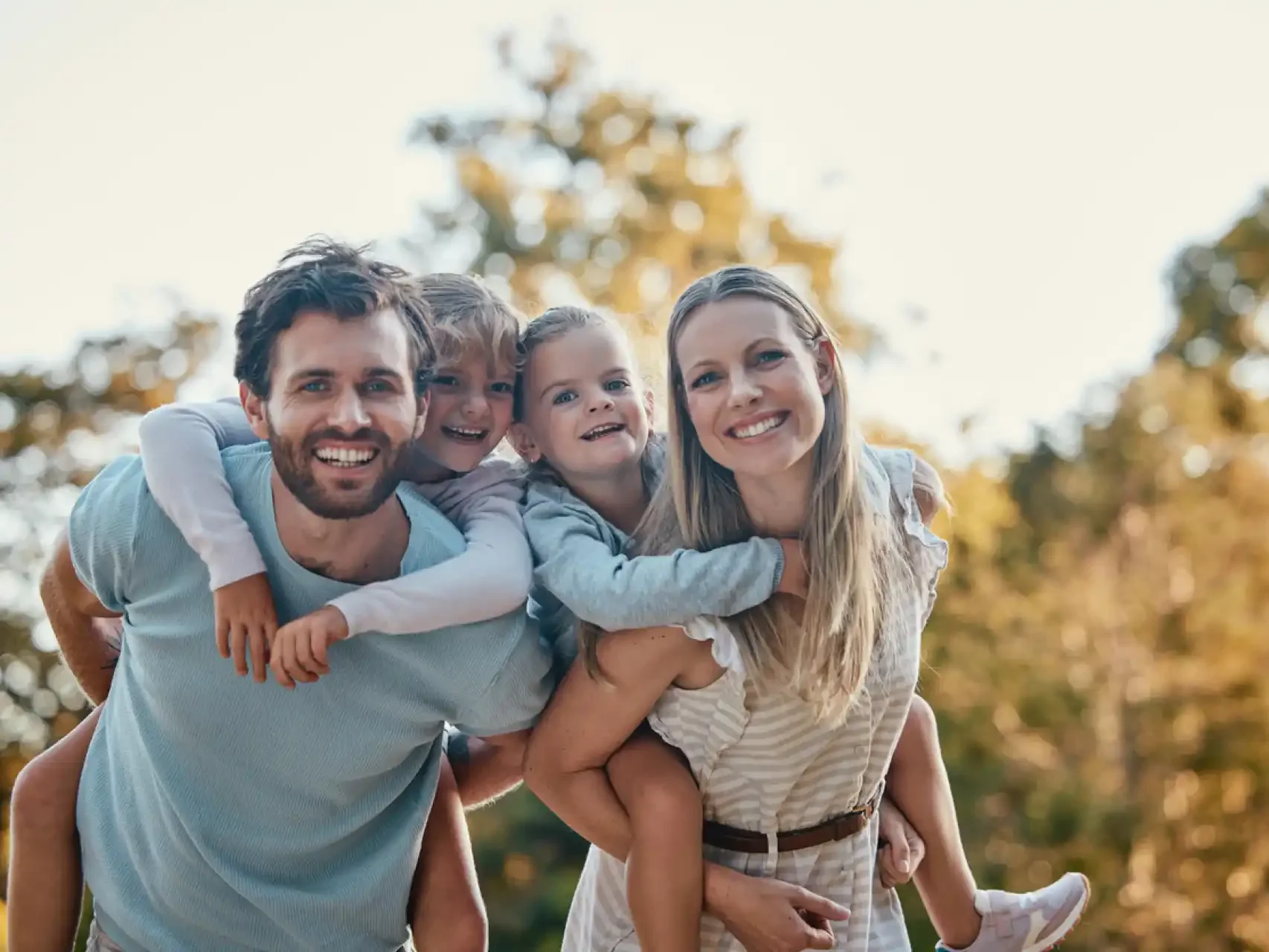 Familia feliz en parque con niños en brazos y adultos sonriendo bajo árboles.