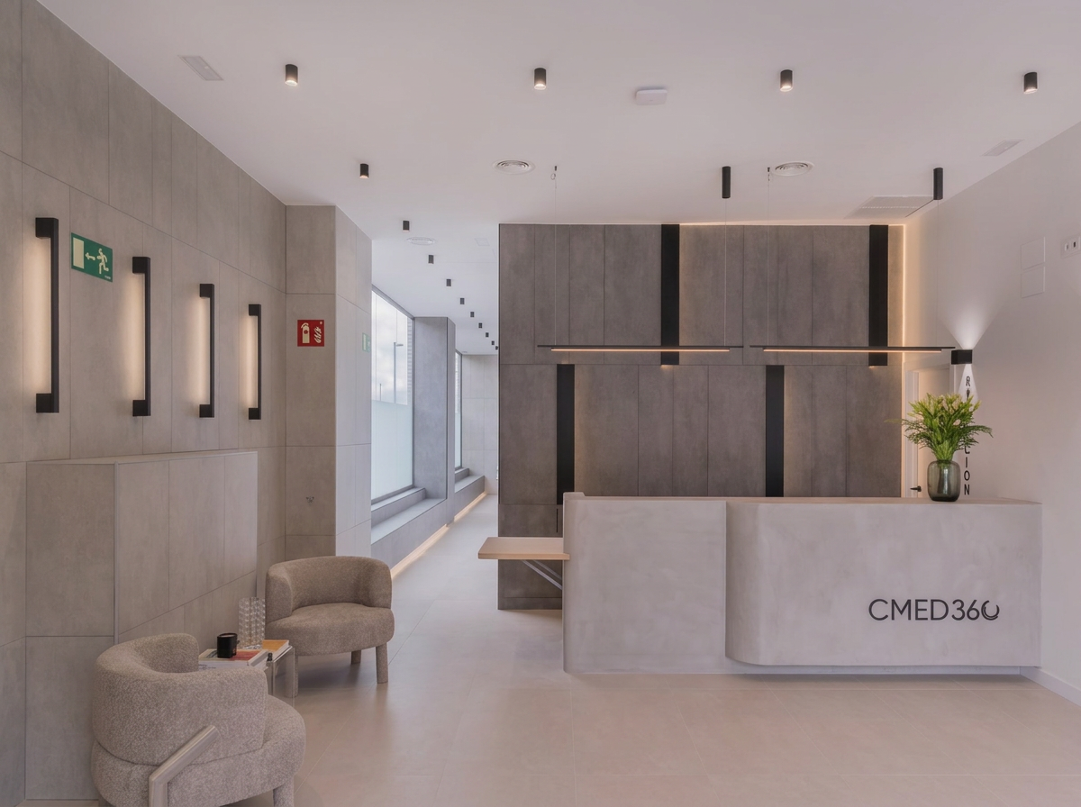 Recepción moderna en un consultorio médico con pared gris, planta en una esquina y sillón beige, cartel verde de salida de emergencia y un cartel rojo de extintor