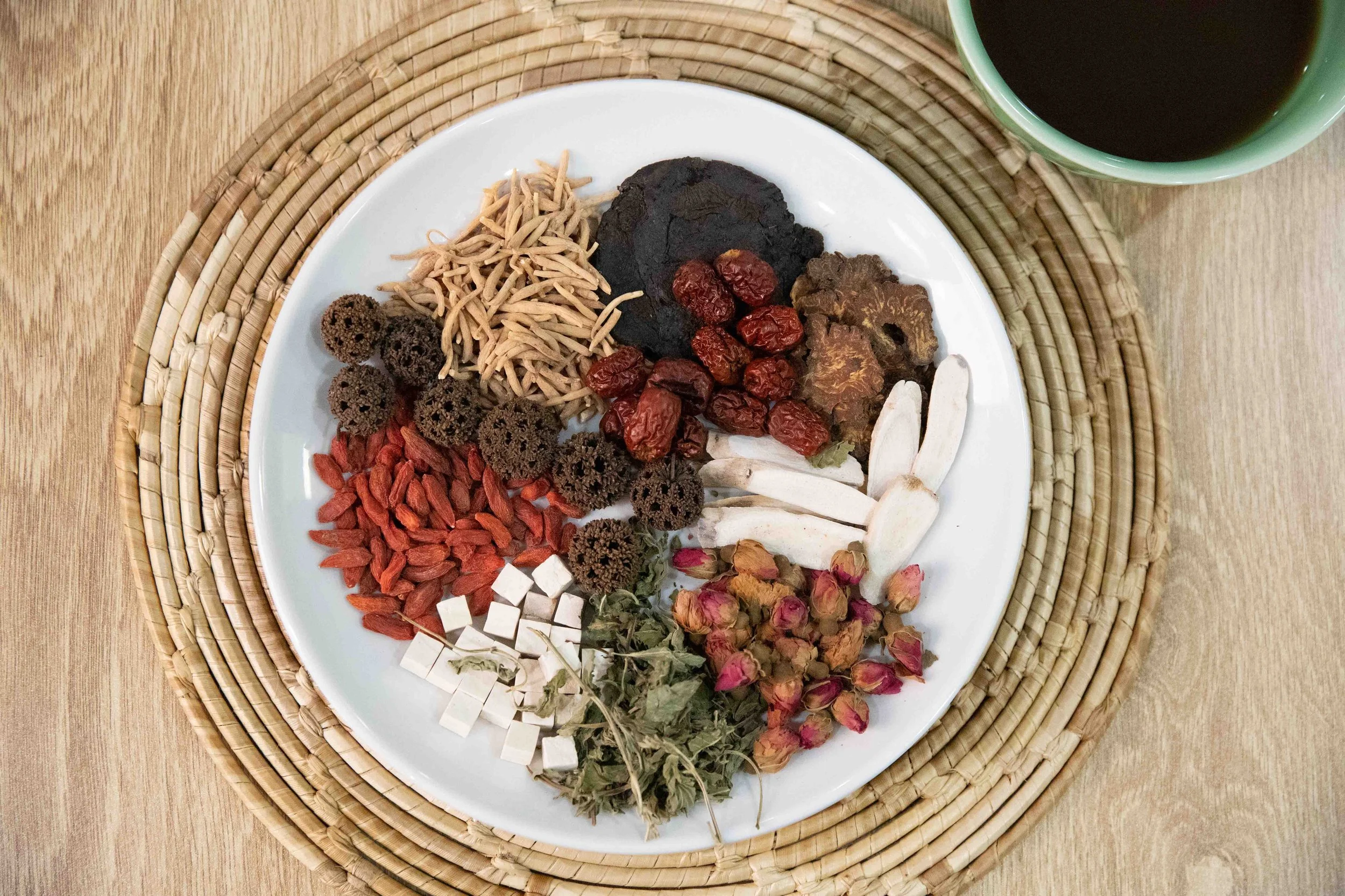 Assorted traditional Chinese medicine ingredients on a white plate, including dried herbs, roots, berries, and flowers, placed on a woven circular mat on a wooden table.