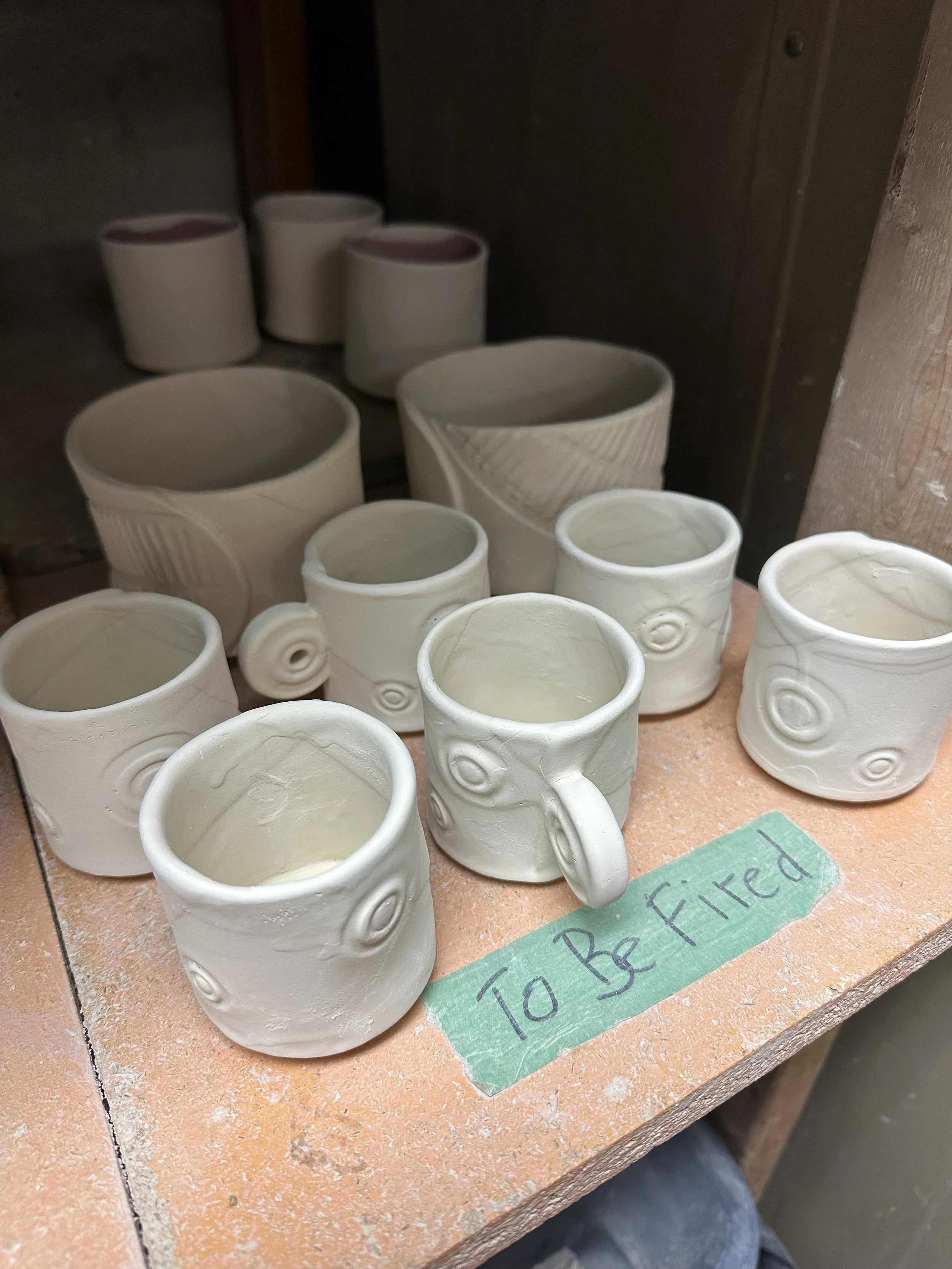 A collection of unfinished white ceramic cups and vases on a wooden shelf, with a green tape label that says 'To Be Fired'.