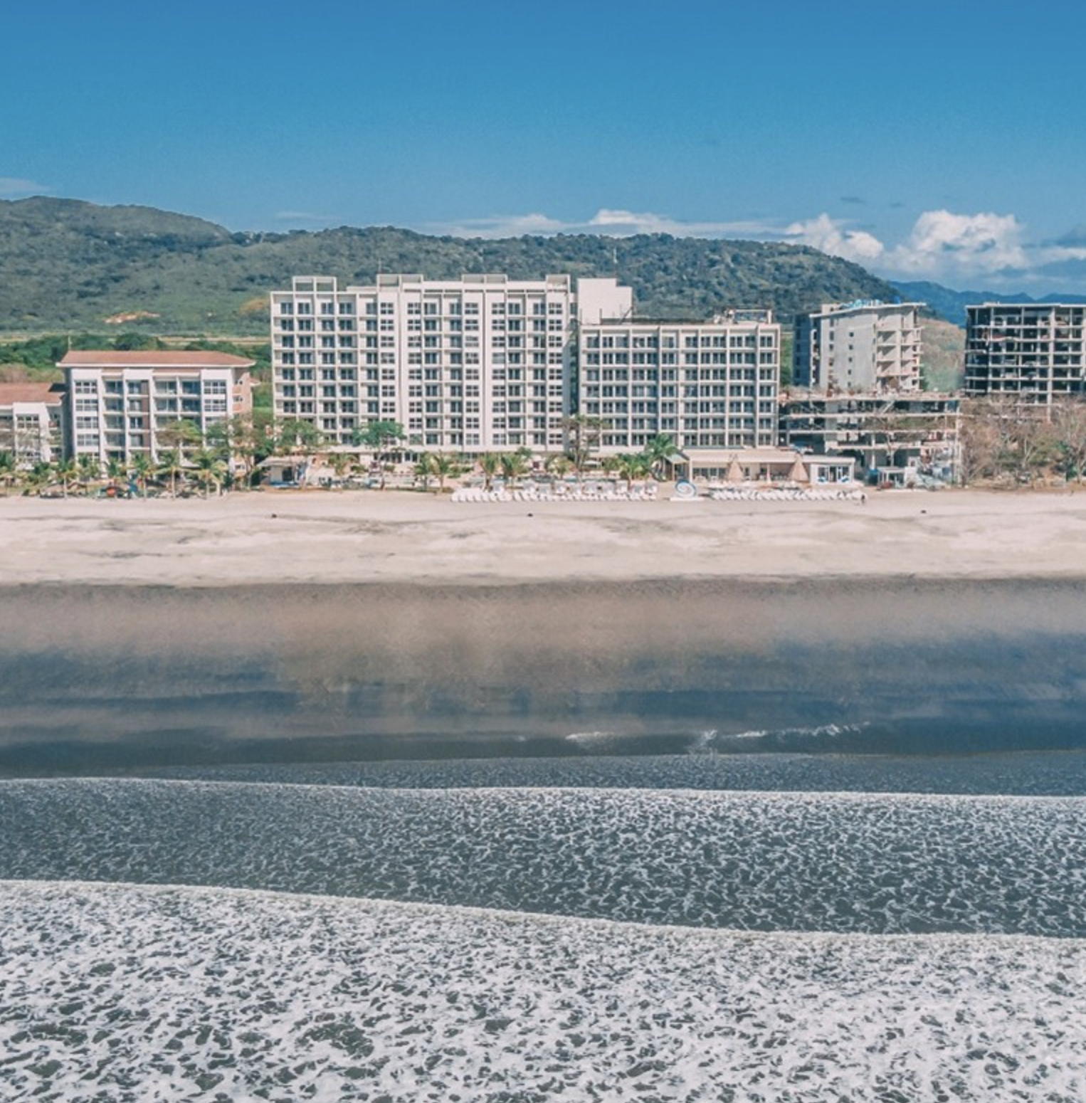 View of beach with sand and waves in the foreground and a row of modern hotel buildings with mountains in the background