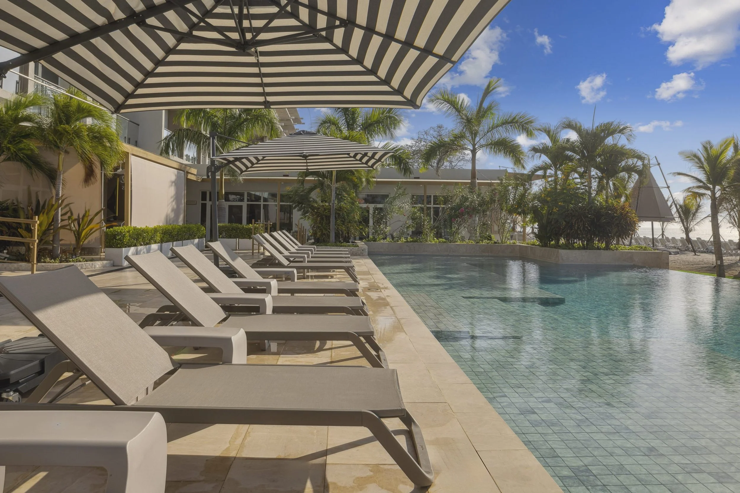 Poolside scene with several lounge chairs under umbrellas, a swimming pool, palm trees, and a building in the background on a sunny day.