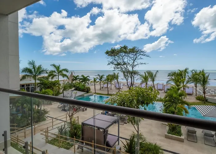 View from a balcony of a resort with palm trees, pool, lounge chairs, and the ocean in the background under a partly cloudy sky.
