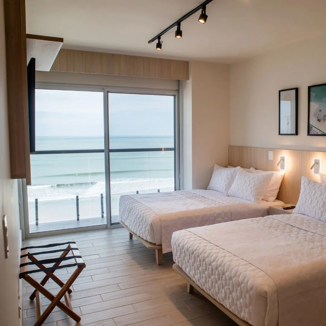 Hotel room with two beds, large sliding glass door showing ocean view, framed artwork, and modern lighting.
