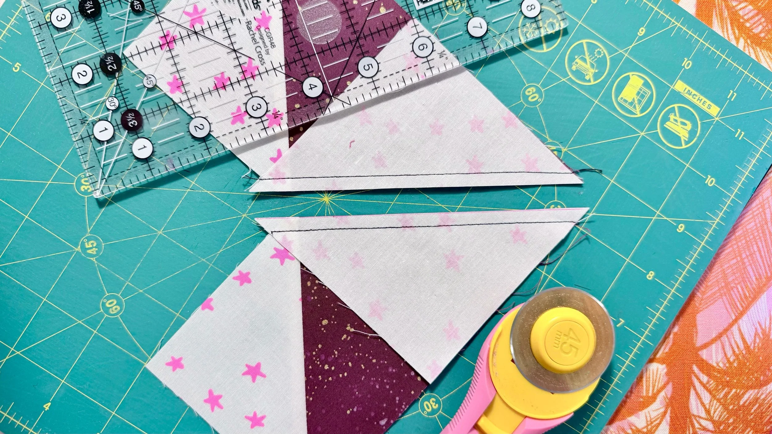 A partially finished maroon and white and pink quilt block cut into 2 mirror image pieces, pictured with a pink rotary cutter and a ruler, on a green cutting mat against a pink and orange background