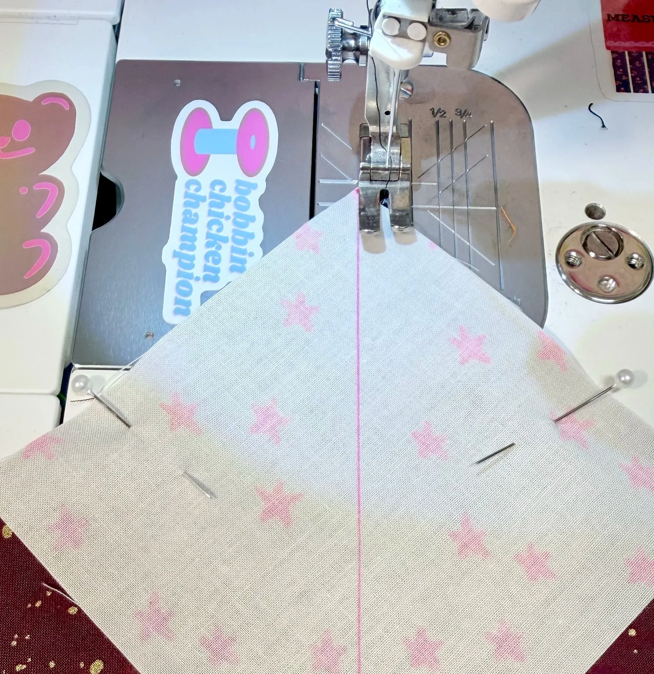 At a sewing machine, a white piece of fabric with a pink line from one diagonal to another is sitting under the presser foot of the machine. It is pinned to sparkly maroon fabric underneath it. The drawn line stretches away from origin.