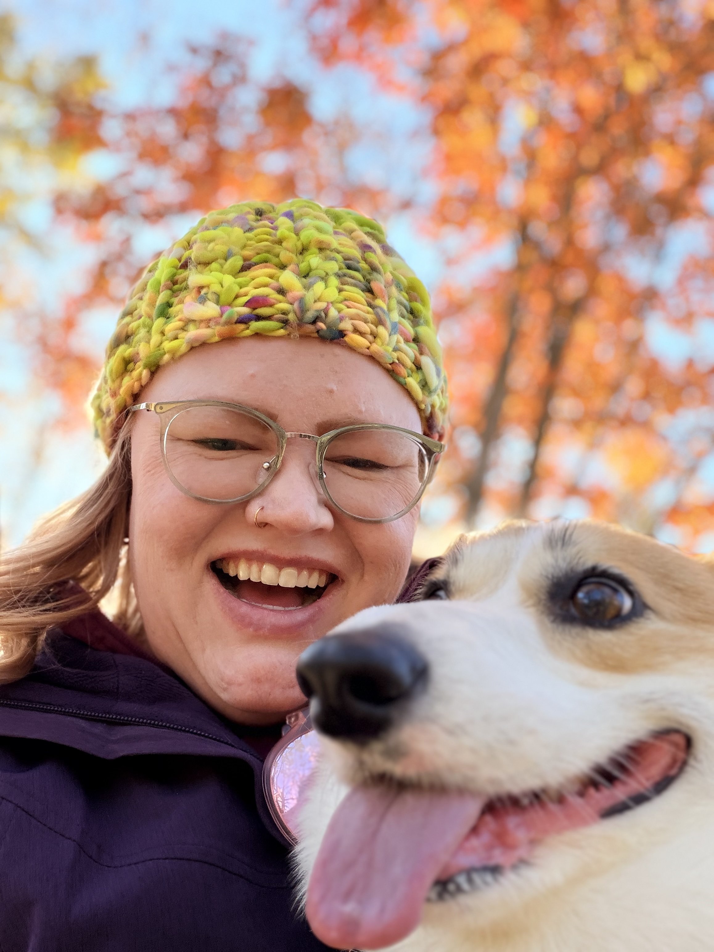 A woman wearing glasses and a colorful knit hat smiling with her orange and white corgi, outdoors with autumn leaves in the background.