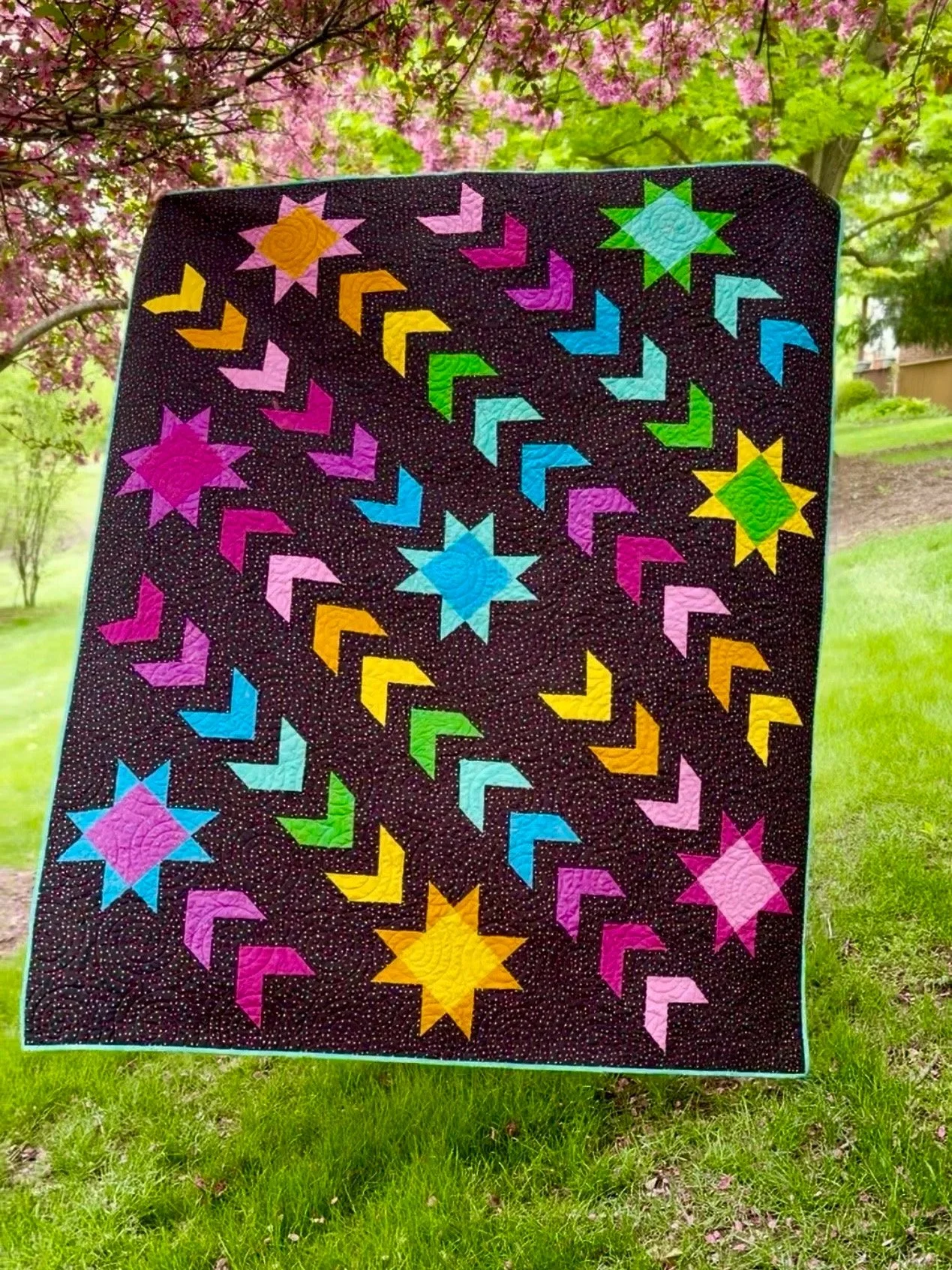 A  quilt with a black background and rainbow colored geometric stars and arrow patterns, hanging outdoors, with pink blossoms and green trees in the background.