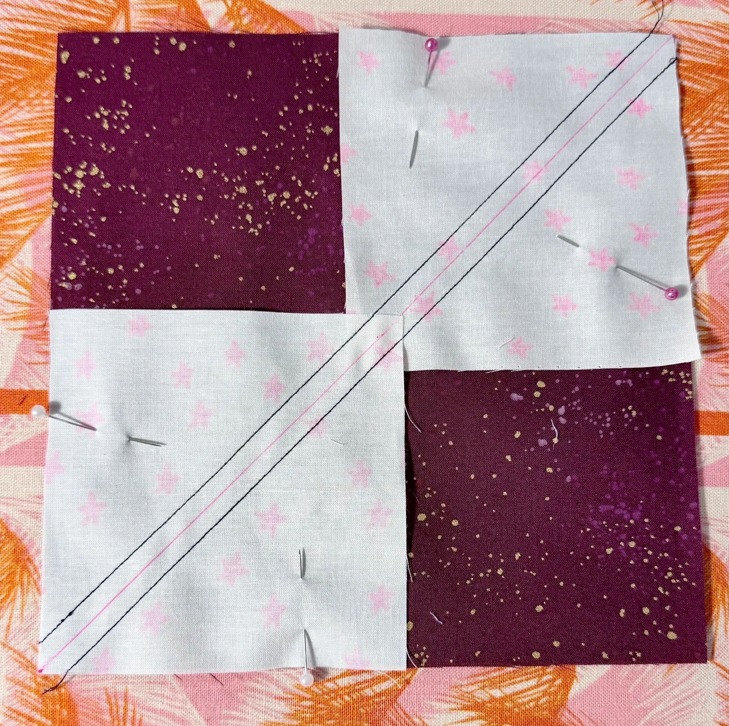 2 squares of white fabric with pink stars are face down, sewn to a face-up maroon fabric square. The small squares line up with opposite corners of the maroon fabric. A pink line follows this diagonal. Black stitching is on both sides of pink line.