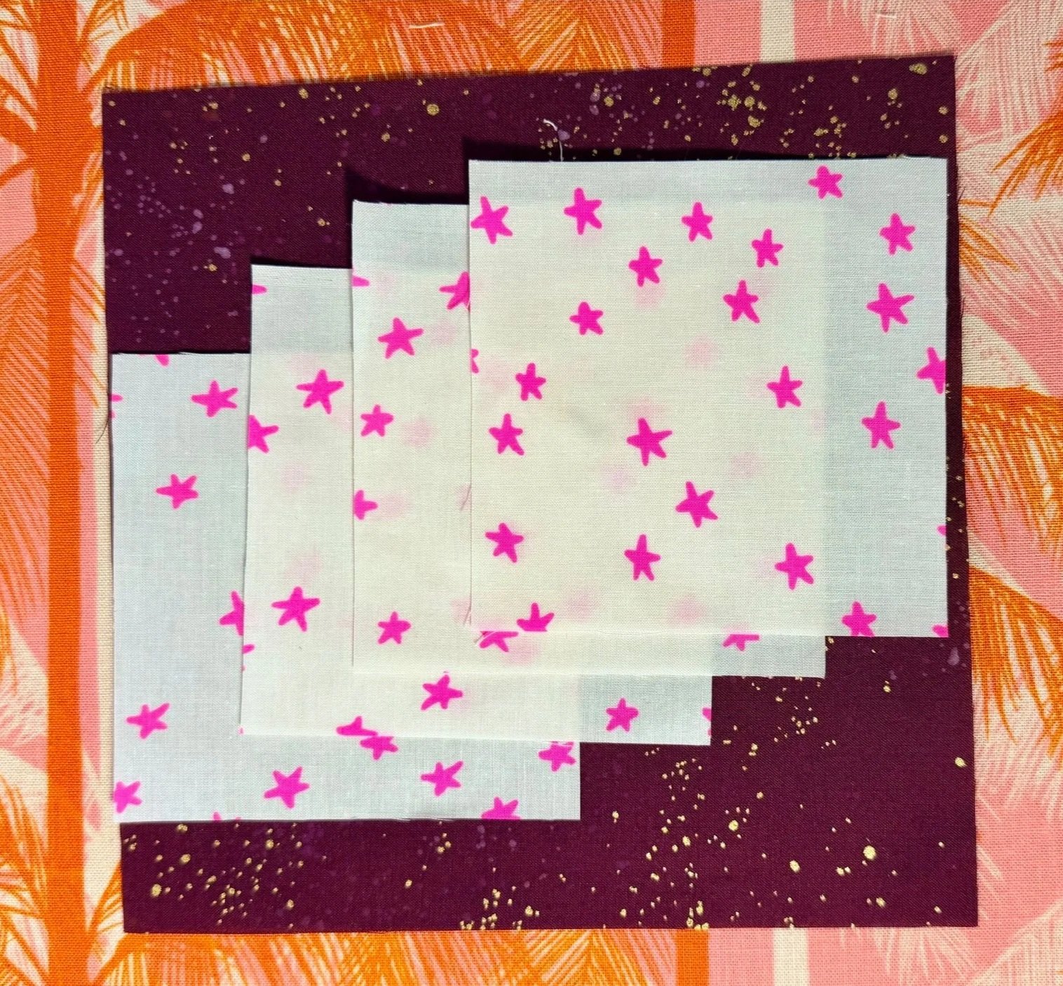 4 fabric squares, 1 larger and sparkly maroon, the smaller 4 with a white background and neon pink stars, are stacked neatly on top of an orange and pink background
