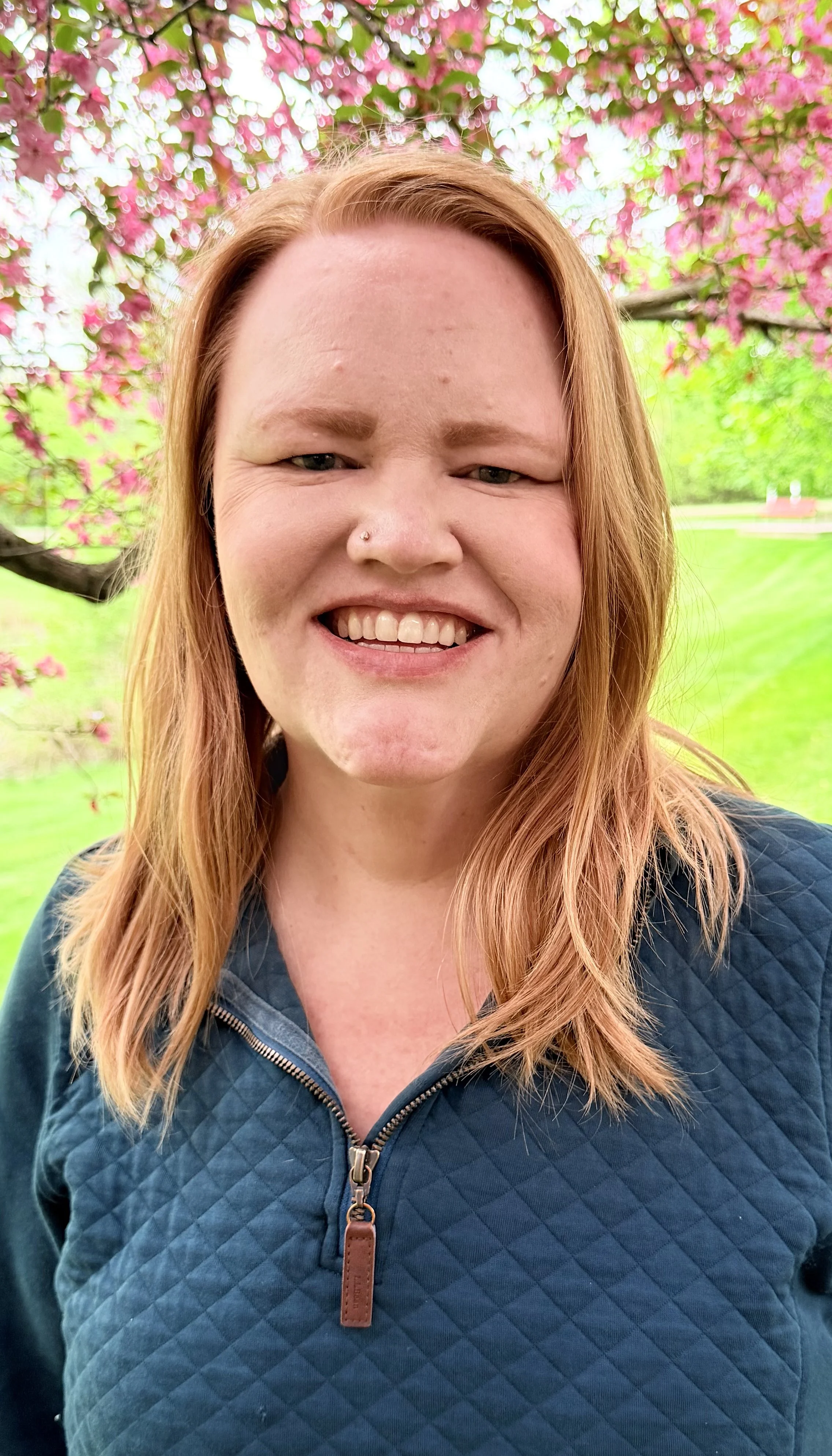 A smiling woman with red hair, a nose piercing, and a blue quilted jacket standing outdoors in front of pink flowering trees and green grass.