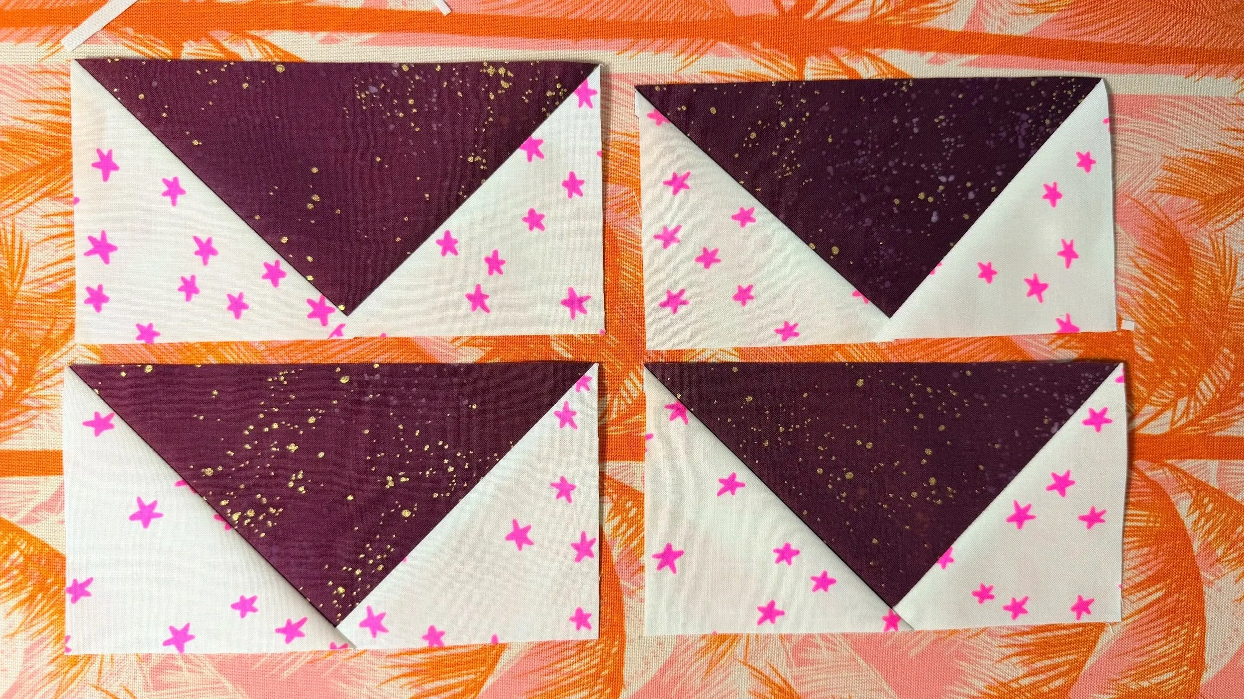4 flying geese quilt blocks on an orange and pink background. Each quilt block has a glittery maroon triangle point down in the middle, with a white triangle with neon pink stars on either side of each maroon triangle.