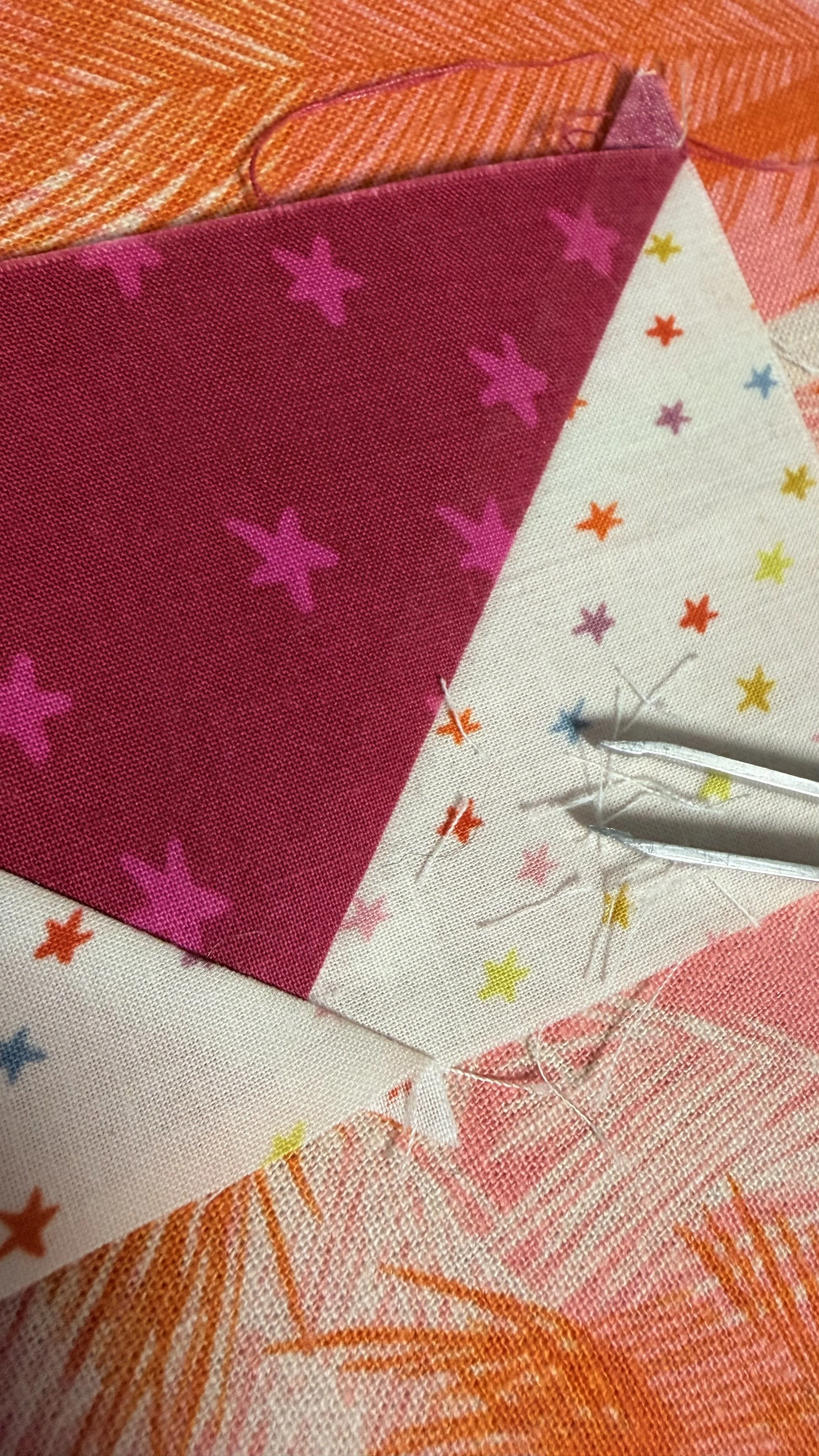 A closeup picture of the point on a flying geese quilt block, illustrating where the corner pieces of fabric come together. Loose threads and a pair of tweezers are sitting on top of the block.