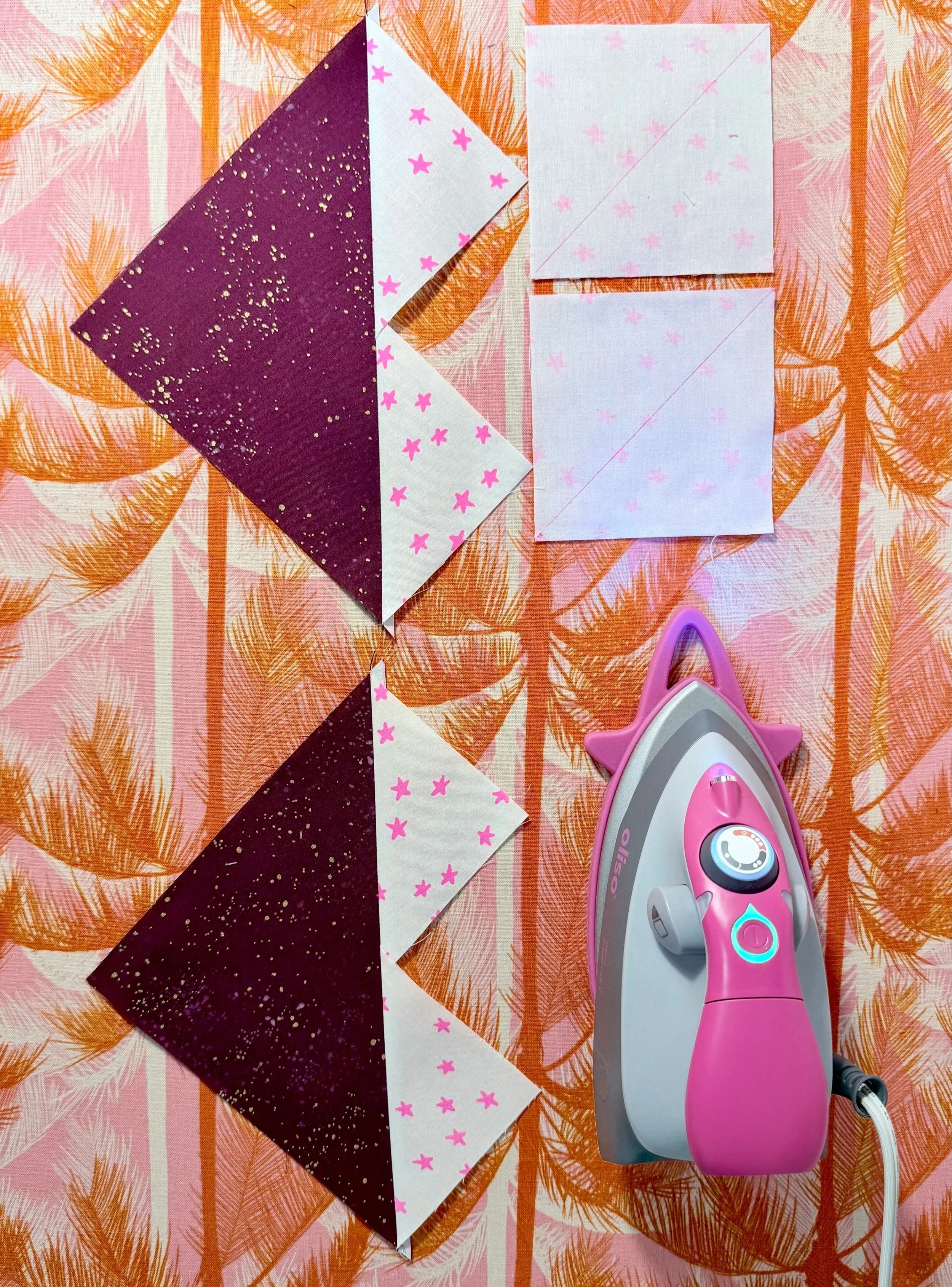 2 heart shaped quilt blocks consisting of sparkly maroon fabric, and white fabric with pink stars, sit on an orange and pink background next to 2 squares of the white fabric and a small pink iron.