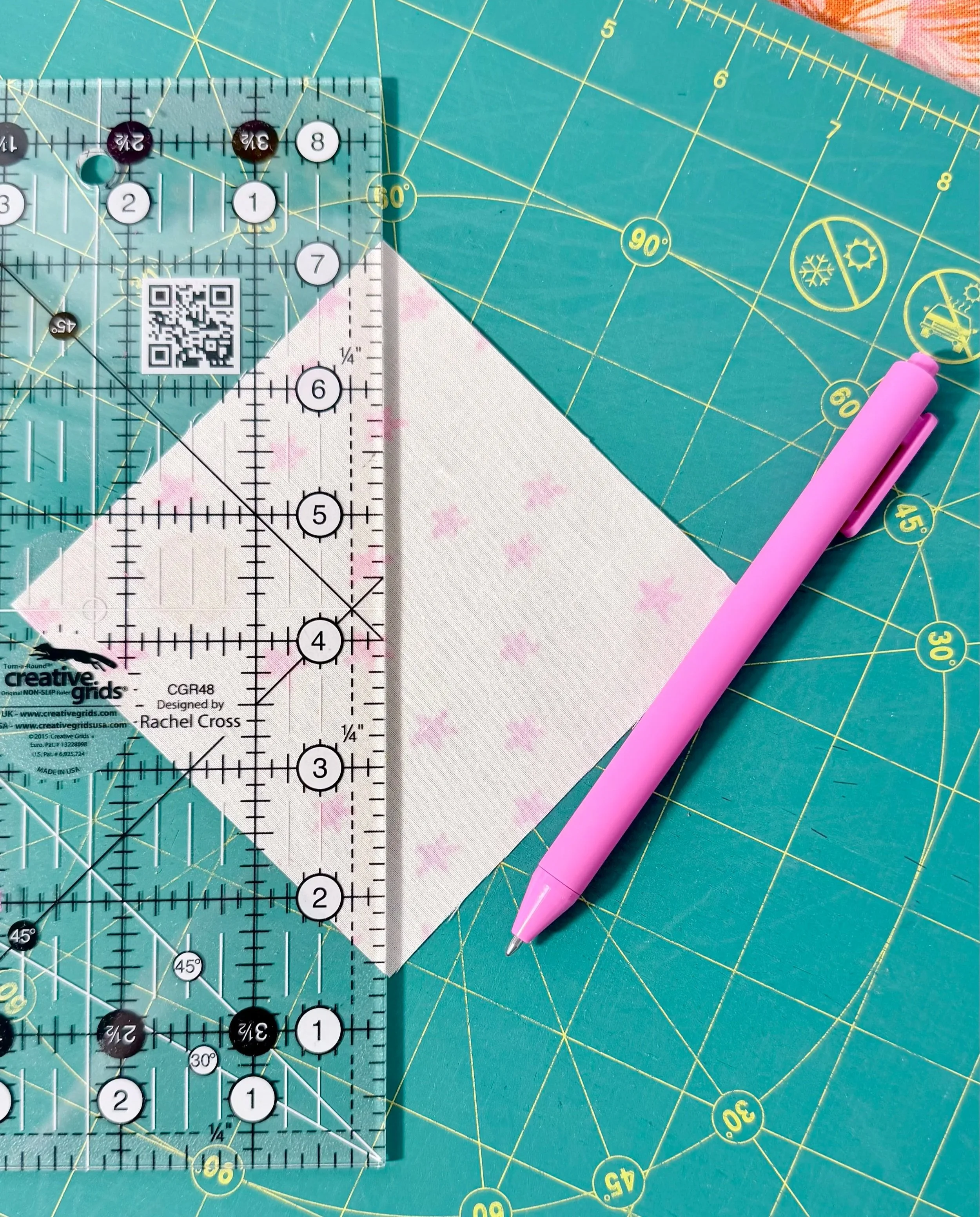 pink pen resting on a green cutting mat with yellow markings, next to a square piece of fabric facing upside down, white with pink stars faintly visible; a clear acrylic ryler with black markings goes from corner to corner on the fabric square