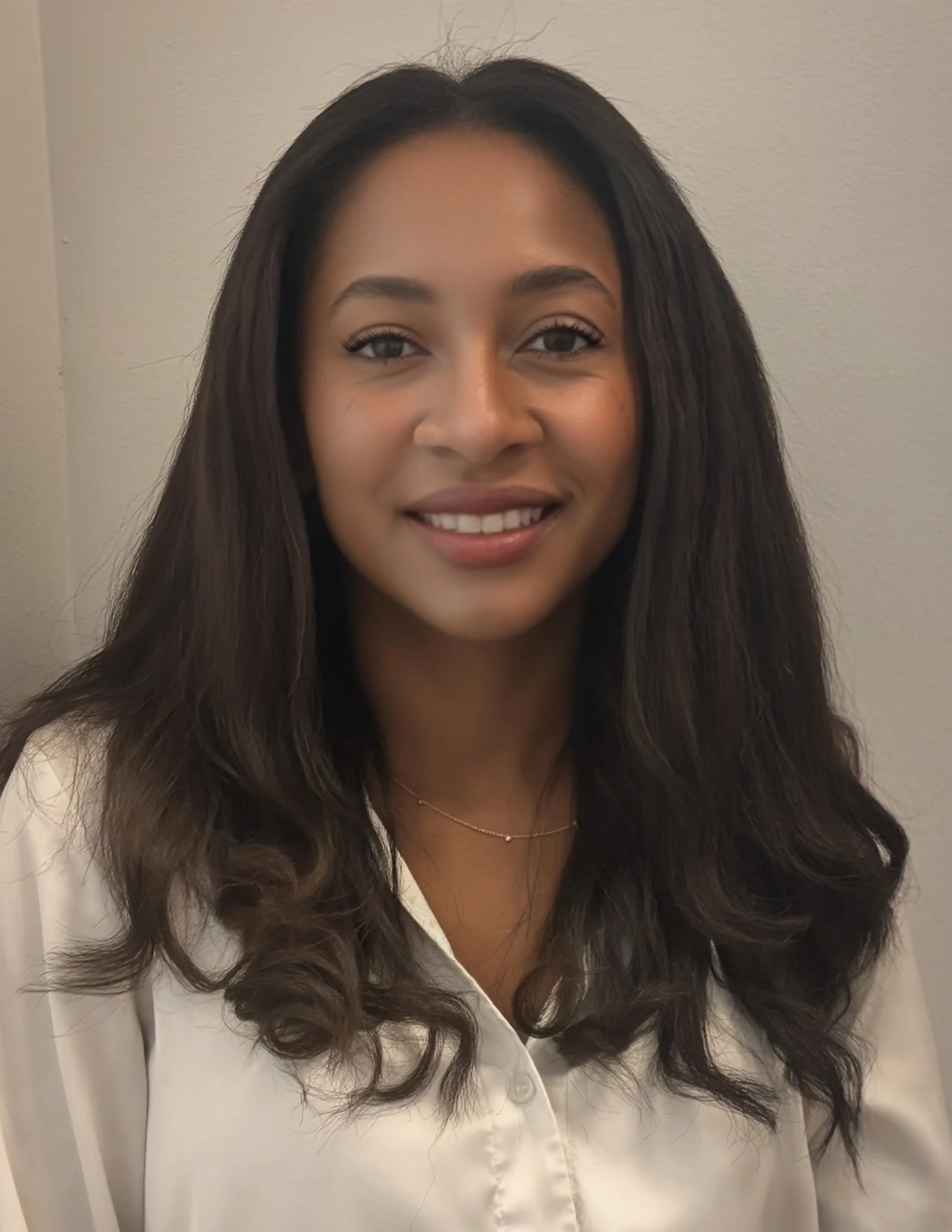 A woman with long dark hair wearing a white top, smiling, standing against a plain wall.
