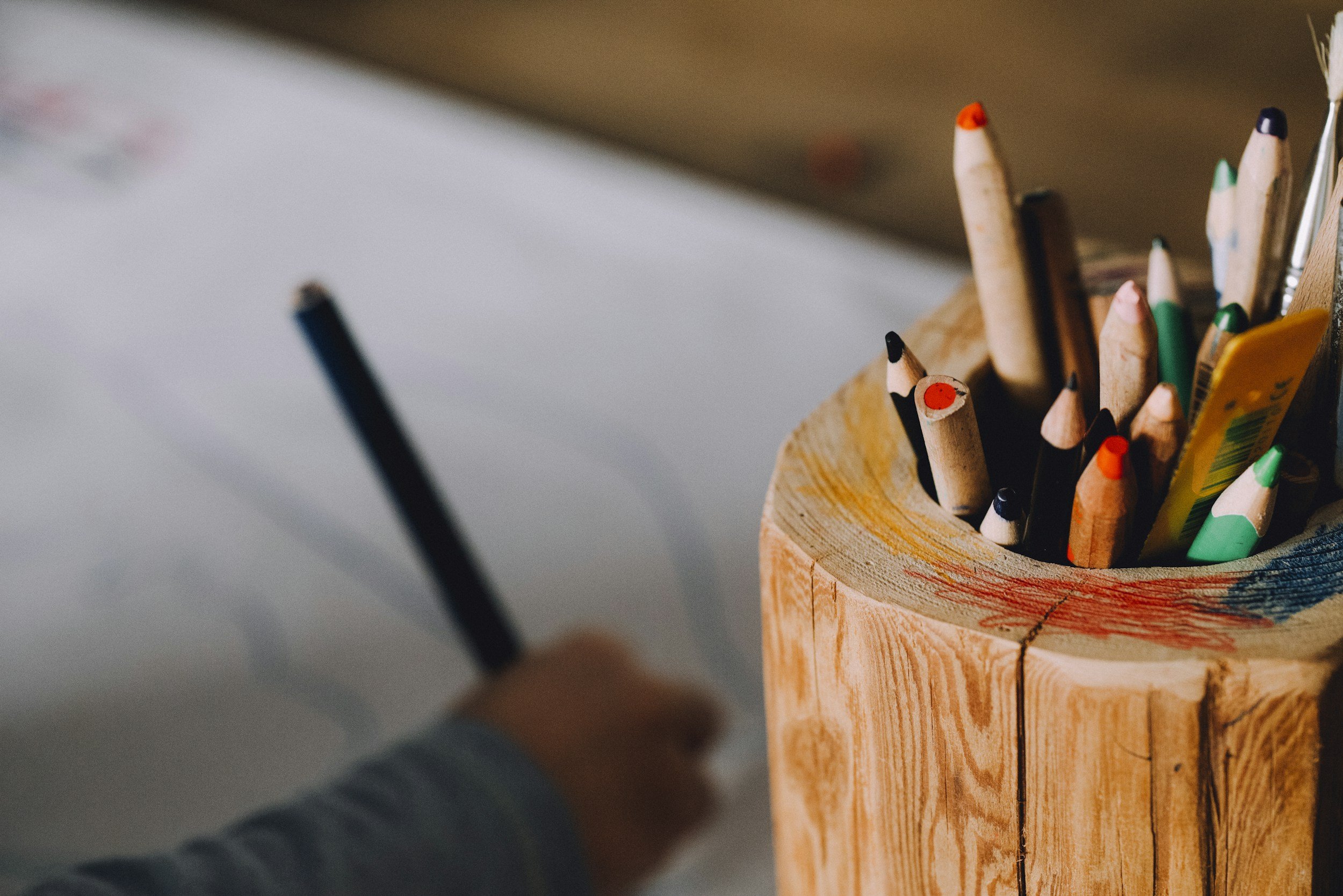Close-up of colored pencils in a wooden holder, with a blurred hand holding a pencil in the background.