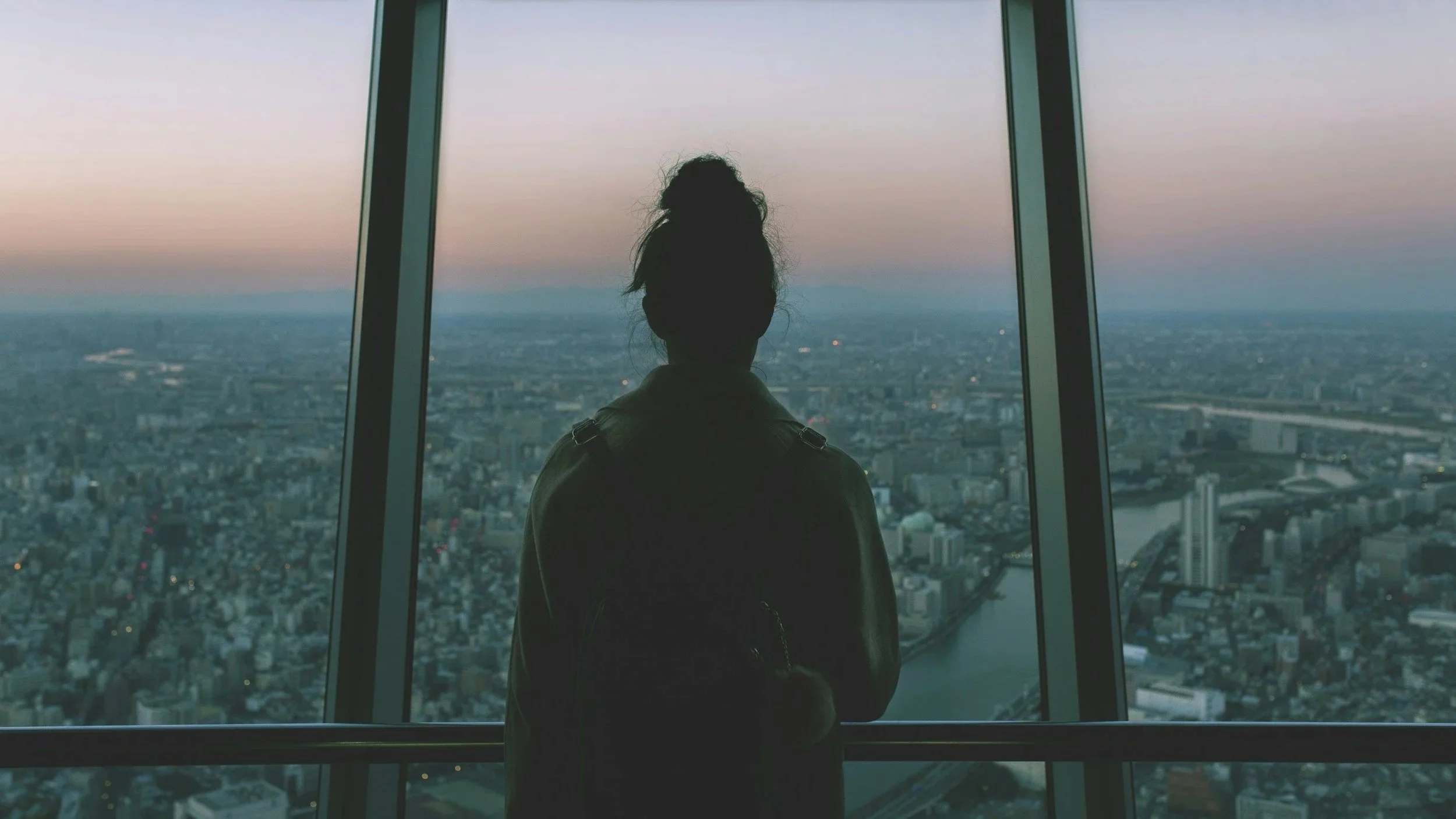 Parent sitting alone and looking out over a city at dusk, representing postpartum therapy for parents feeling overwhelmed.