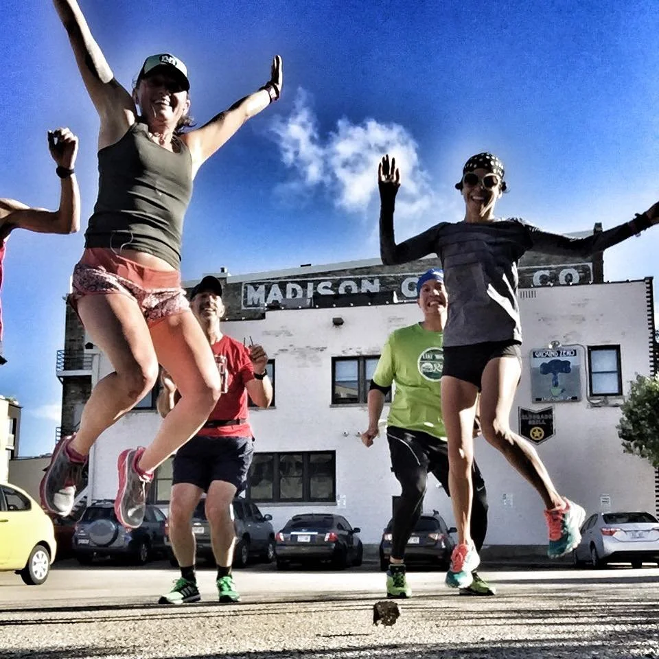 Group of people jumping in front of a building that says "MADISON" on the sign, on a sunny day with a blue sky and some clouds.