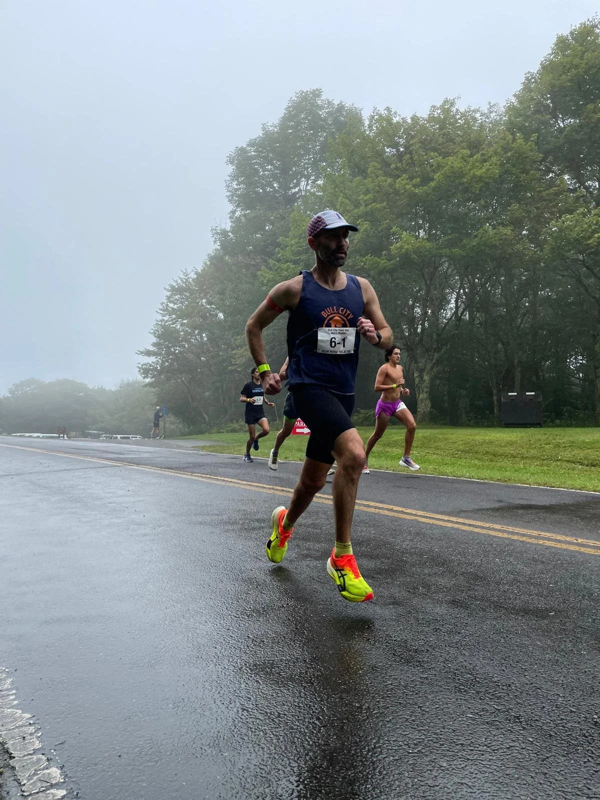 A man running in a race on a wet, foggy road, wearing a blue tank top, black shorts, yellow and pink running shoes, and a baseball cap, with a couple of other runners in the background.