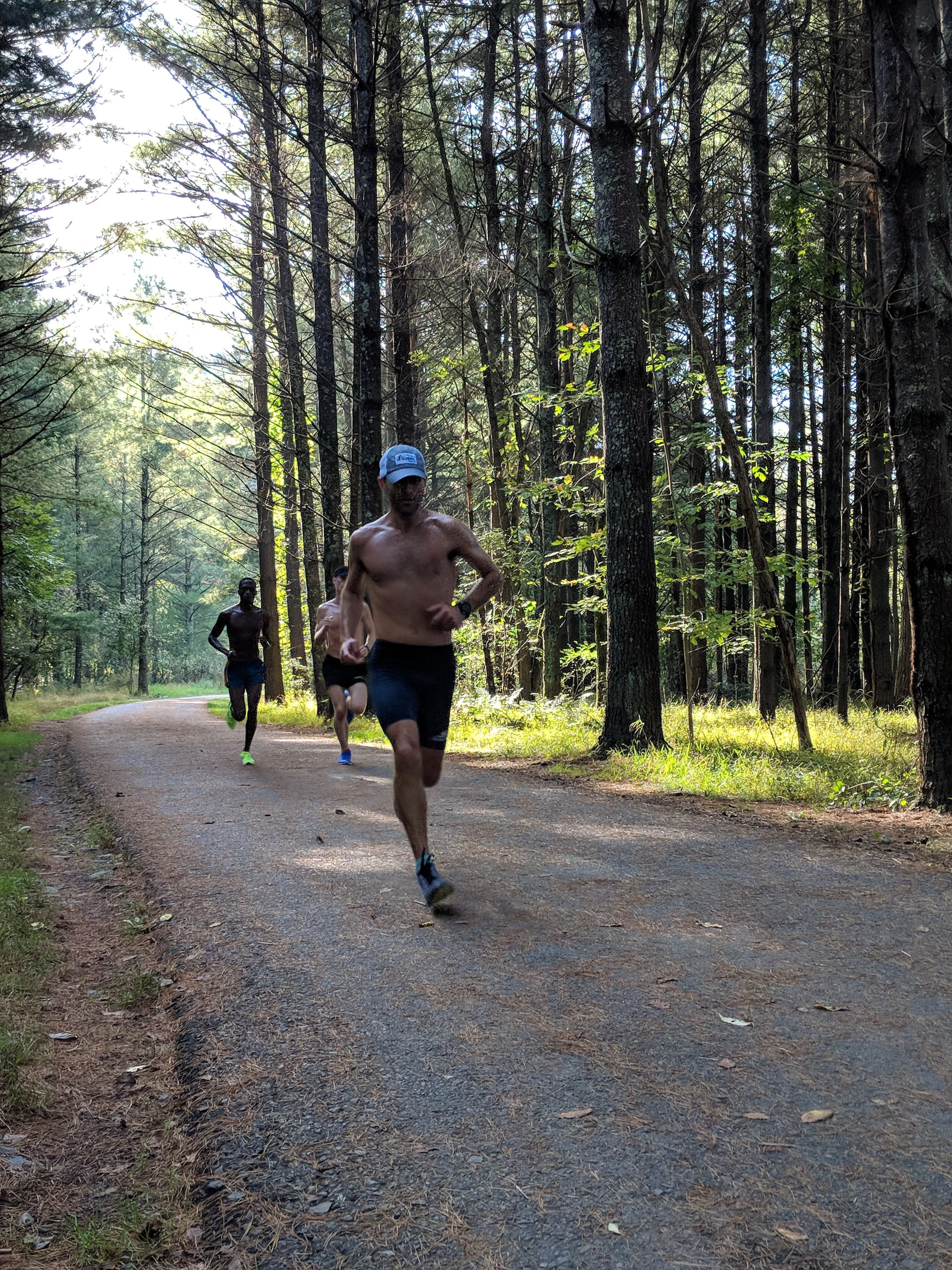 Three men running shirtless on a dirt trail through a forest with tall trees and green foliage.