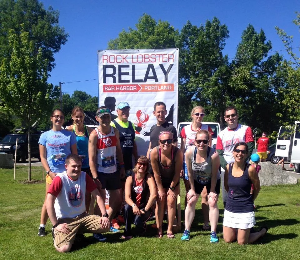 Group of people posing outdoors during a relay race event called Rock Lobster Relay in Portland, near Bar Harbor, with a large sign in the background showing the event name and location.