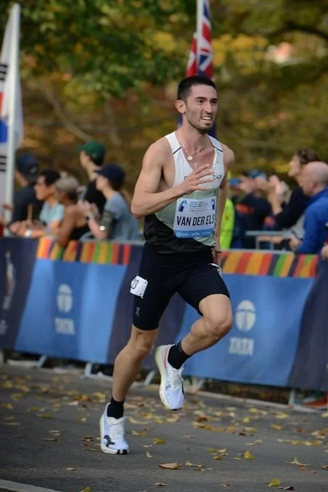 Male marathon runner wearing a white running top and black shorts running with a crowd behind barriers and autumn trees in the background.