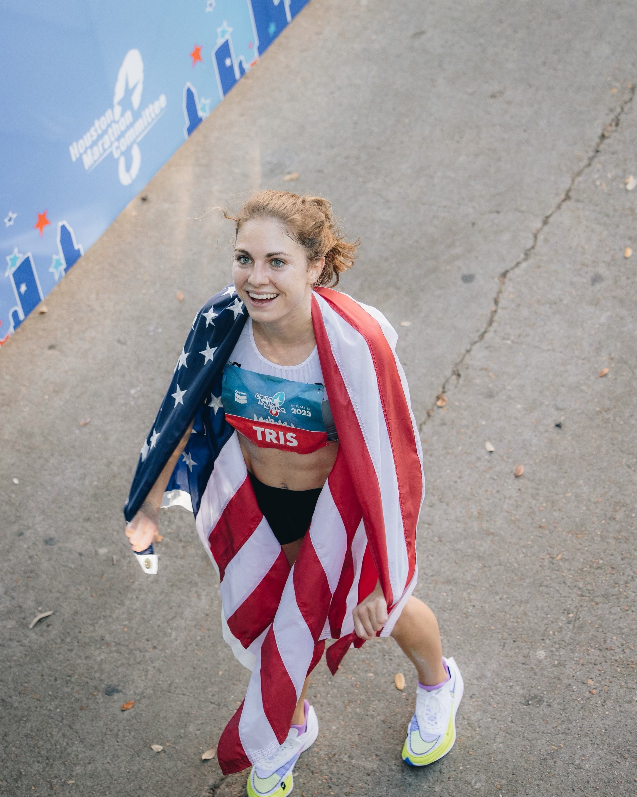 A female athlete smiling while running, draped in an American flag after a race, wearing a bib with 'TRIS' and '2023' on it.