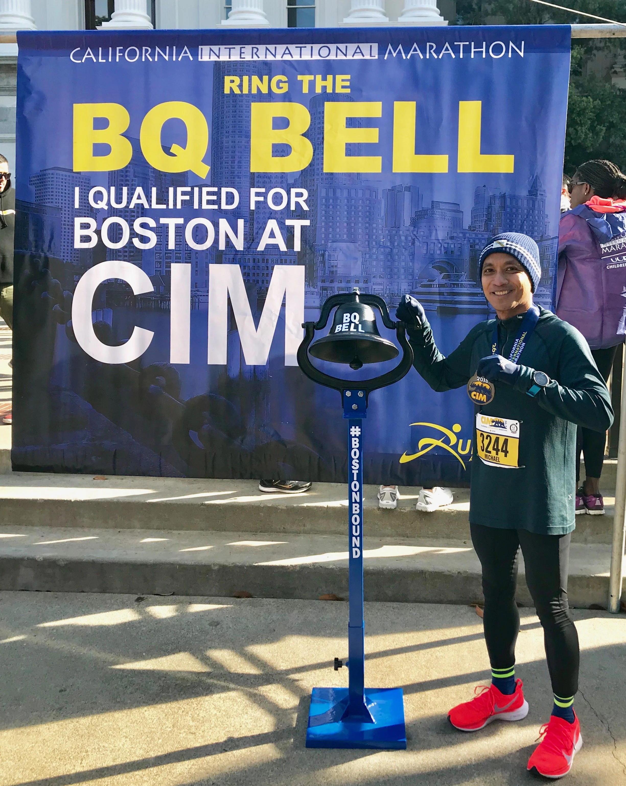 Man in running gear standing next to a bell with an International Marathon banner in the background. The banner reads 'California International Marathon, Ring the BQ Bell, I qualified for Boston at CIM' with cityscape images.
