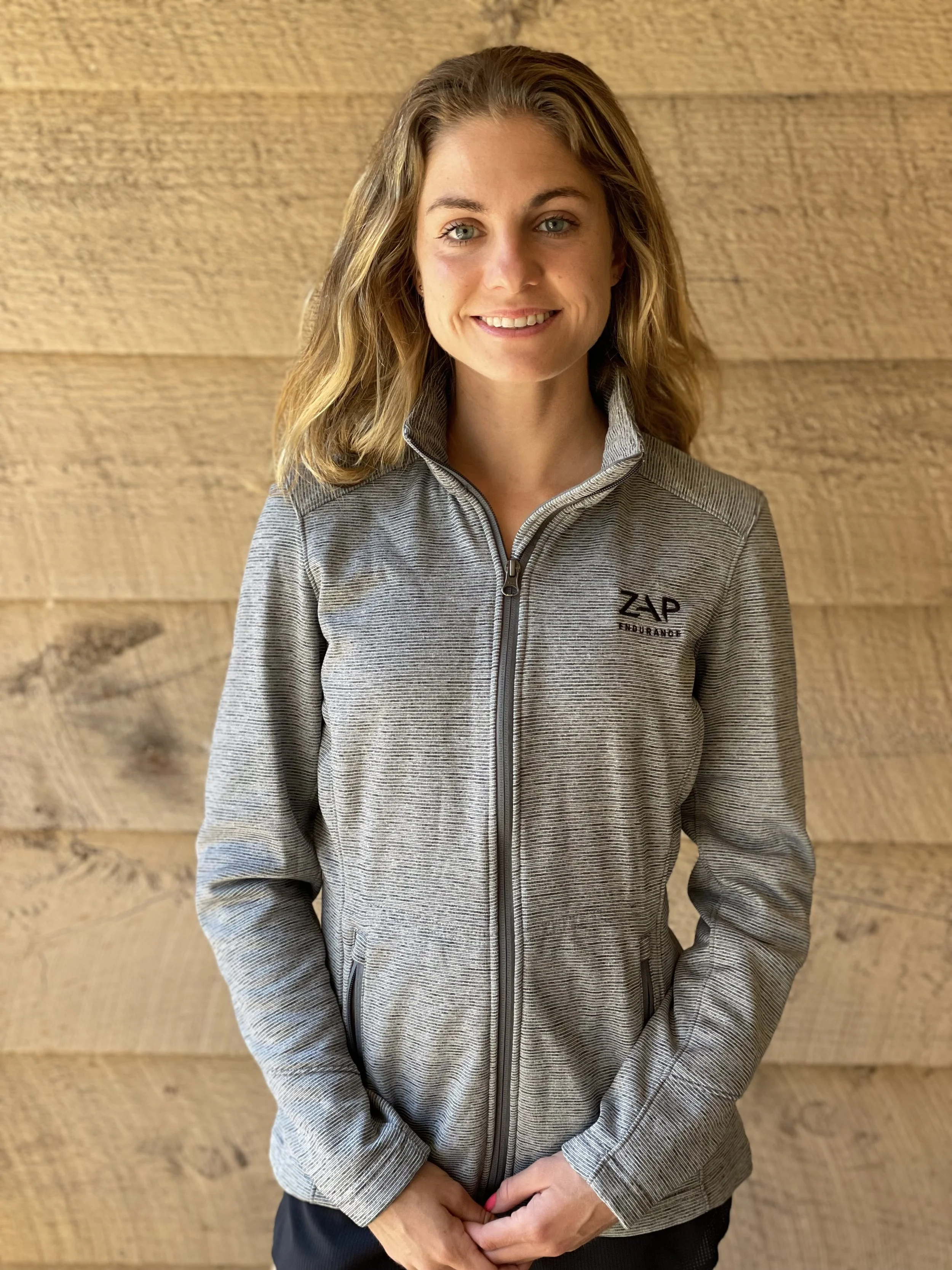 Smiling woman with wavy blonde hair wearing a gray zip-up jacket standing against a wooden wall background.