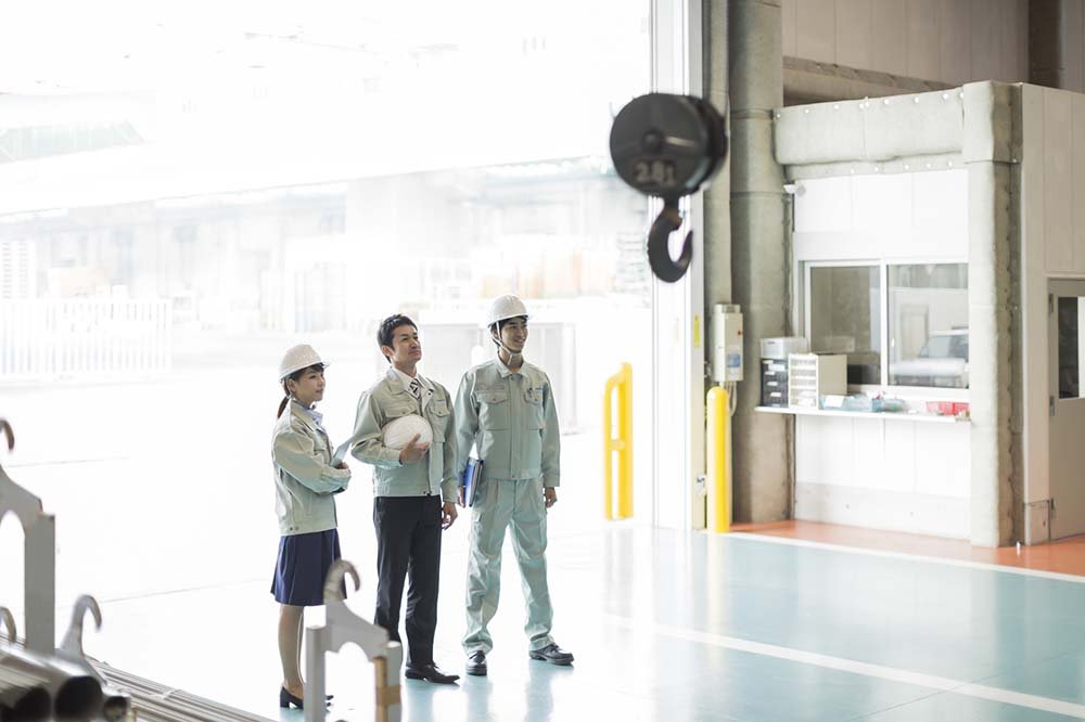 Three people, two men and a woman, standing inside a building near a large open door, all wearing safety helmets and work uniforms.