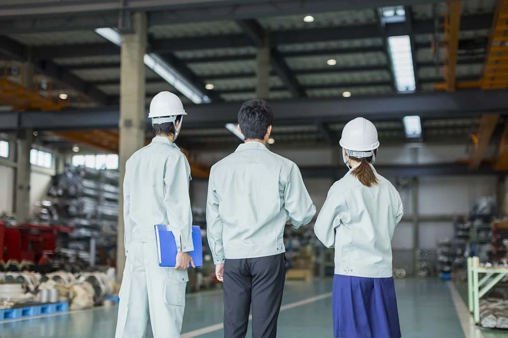 Three workers in safety helmets and work uniforms in an industrial warehouse, with shelves of supplies and equipment in the background.