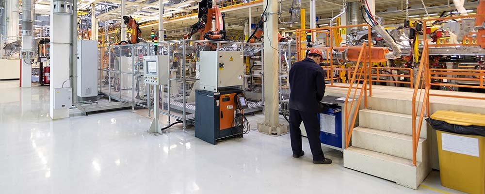 Factory interior with safety barriers, robotic arms, and a worker operating a control panel near stairs in an industrial setting.