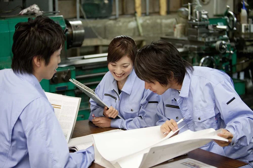 Three people in blue work uniforms sitting at a table with blueprints and documents, engaged in a discussion in an industrial workshop with machinery in the background.