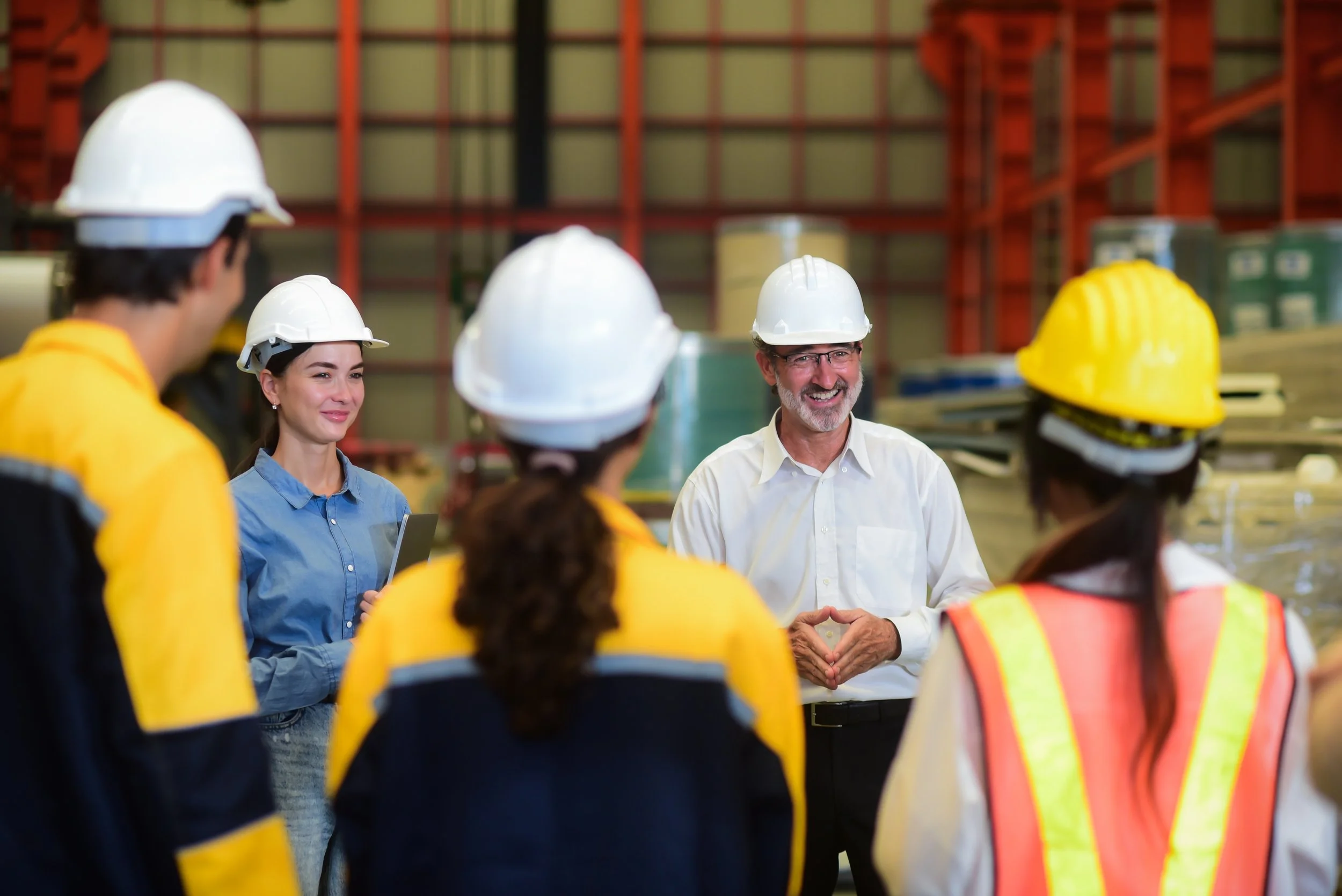 A group of industrial workers wearing safety helmets listening to a man speaking in a warehouse.