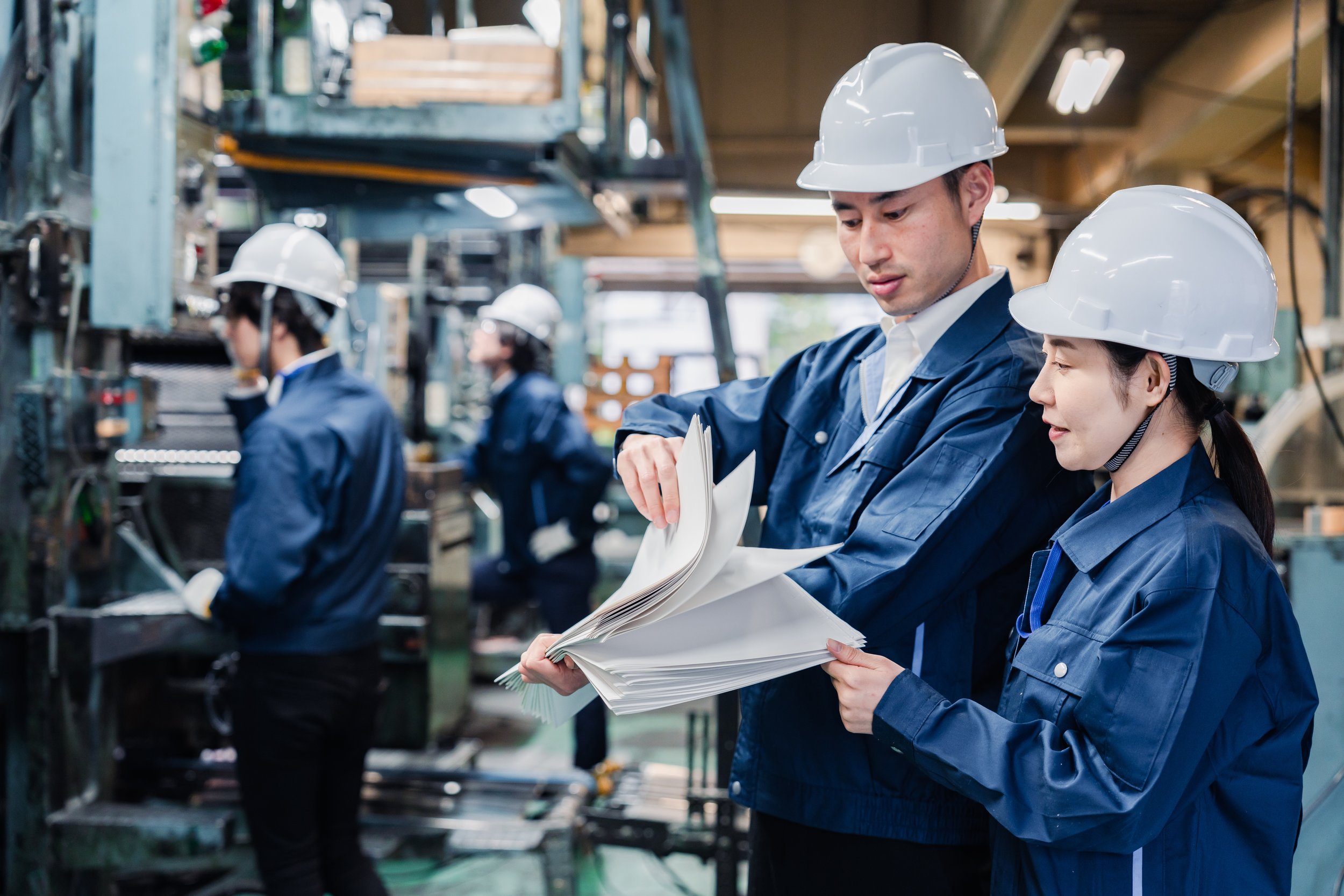 Workers in a factory wearing blue uniforms and white safety helmets, reviewing documents, with machinery in the background.