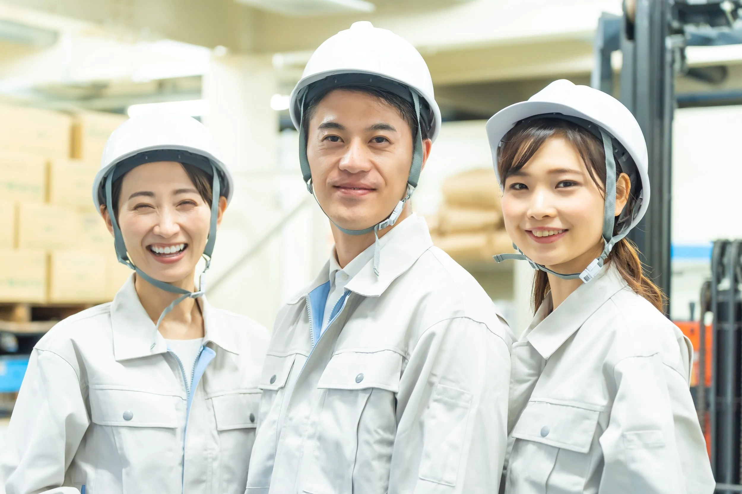 Three factory workers wearing white helmets and uniforms standing in a warehouse, smiling at the camera.
