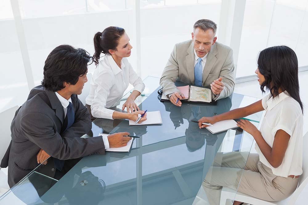 Business meeting with five people sitting around a glass table in a well-lit office, engaged in discussion.