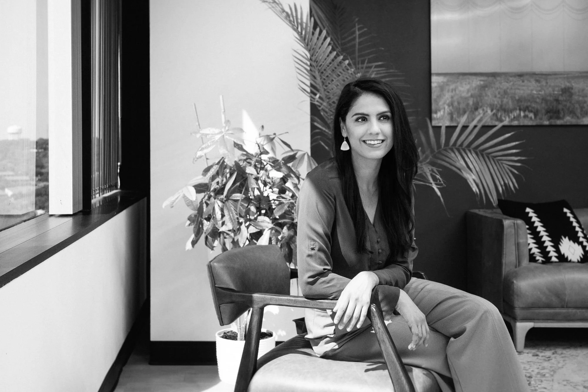 A smiling woman with long dark hair, wearing earrings and a long-sleeve shirt, sitting on a chair in a modern indoor setting with plants, a window, and a sofa in the background.