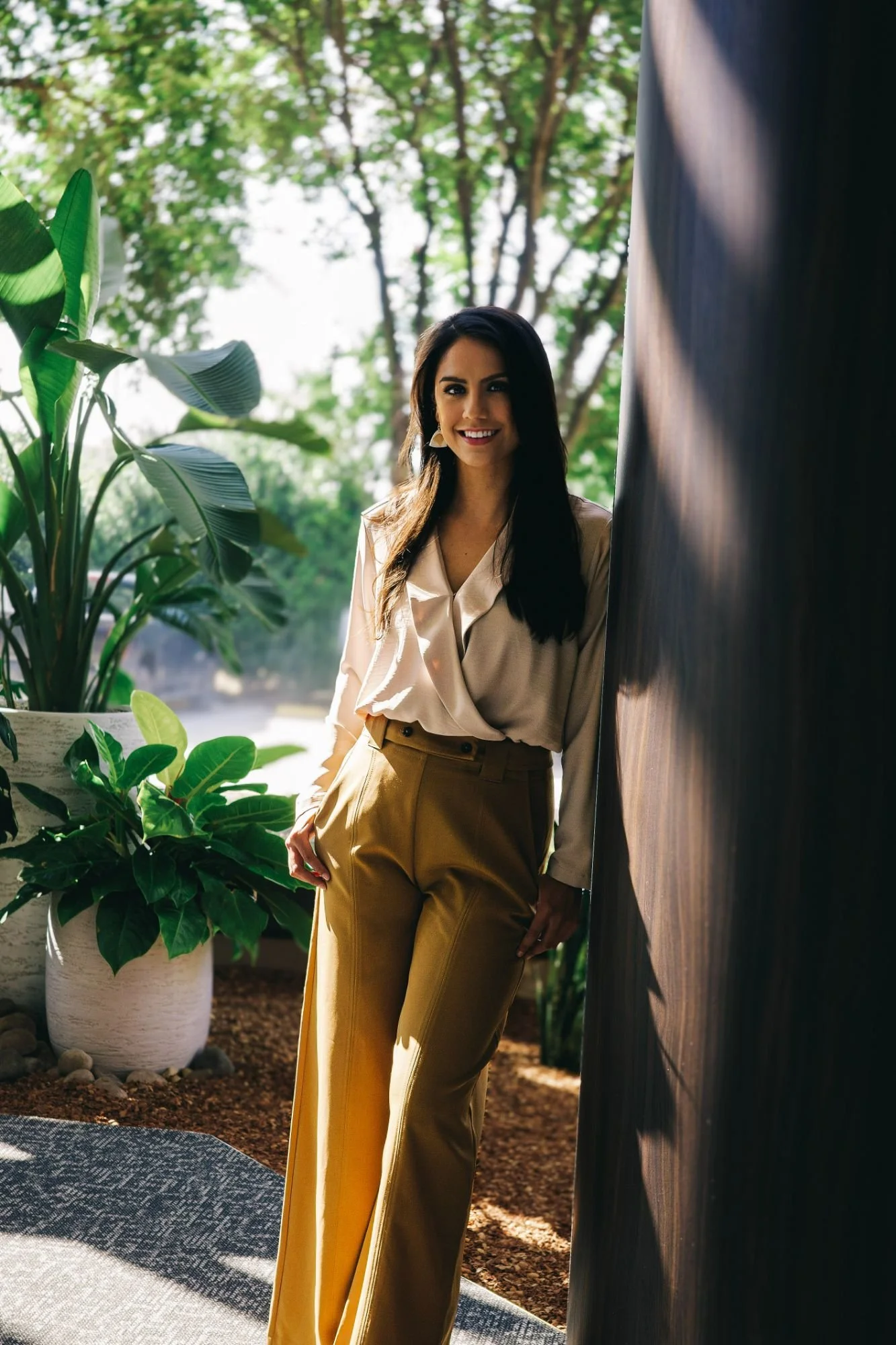 A woman with dark hair, wearing a beige blouse and yellow pants, stands outdoors near a dark wooden wall and green plants, smiling at the camera.