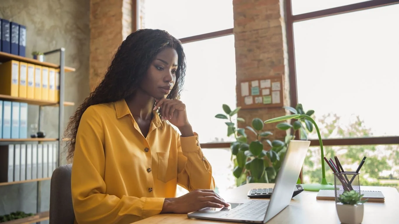 A woman in a yellow shirt working on her laptop at a desk in a bright office with large windows and shelves of binders.