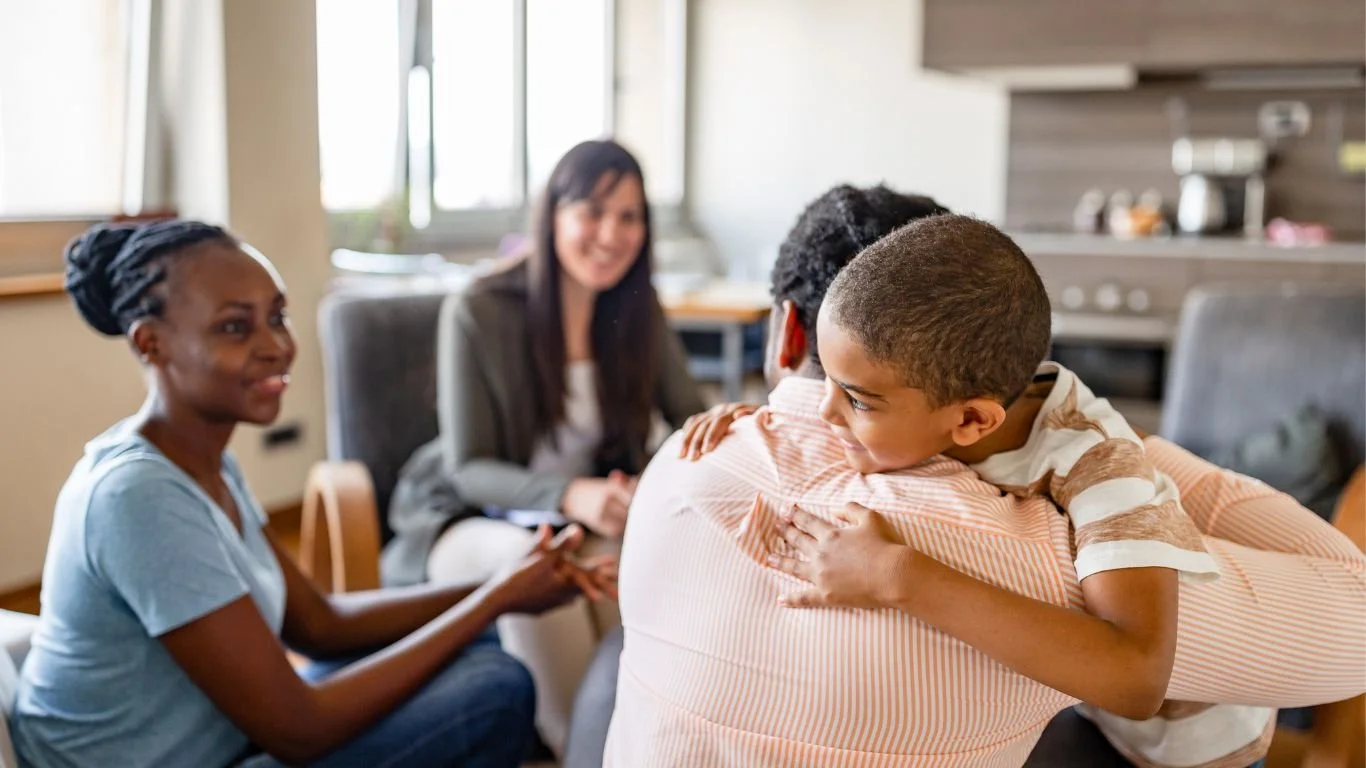 A young boy hugging an adult, smiling, with two women smiling and sitting nearby in a consultation room.