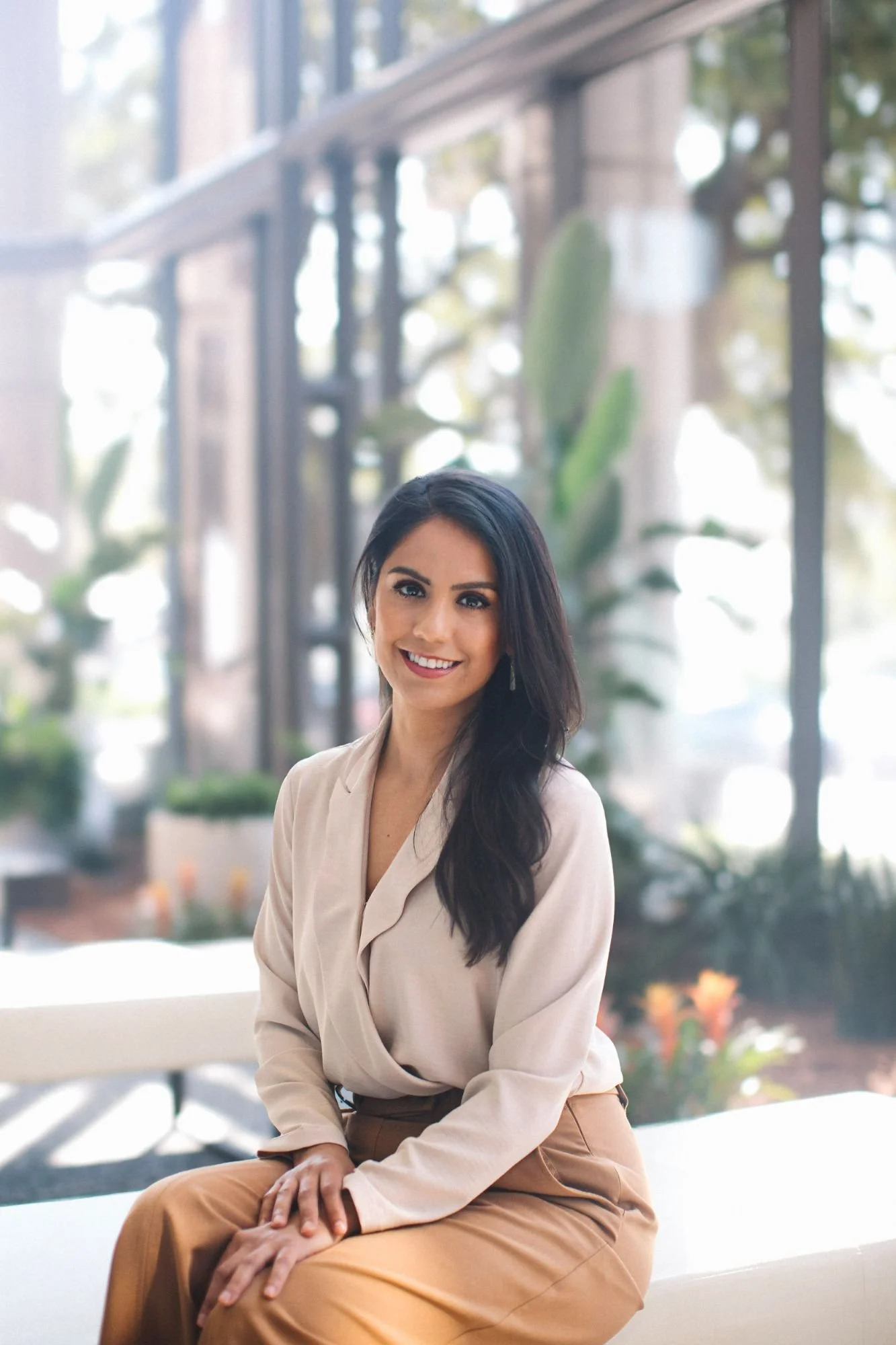 A woman with long black hair, wearing a light beige blouse and tan pants, smiling and sitting on a bench in a well-lit indoor space with large windows and green plants in the background.