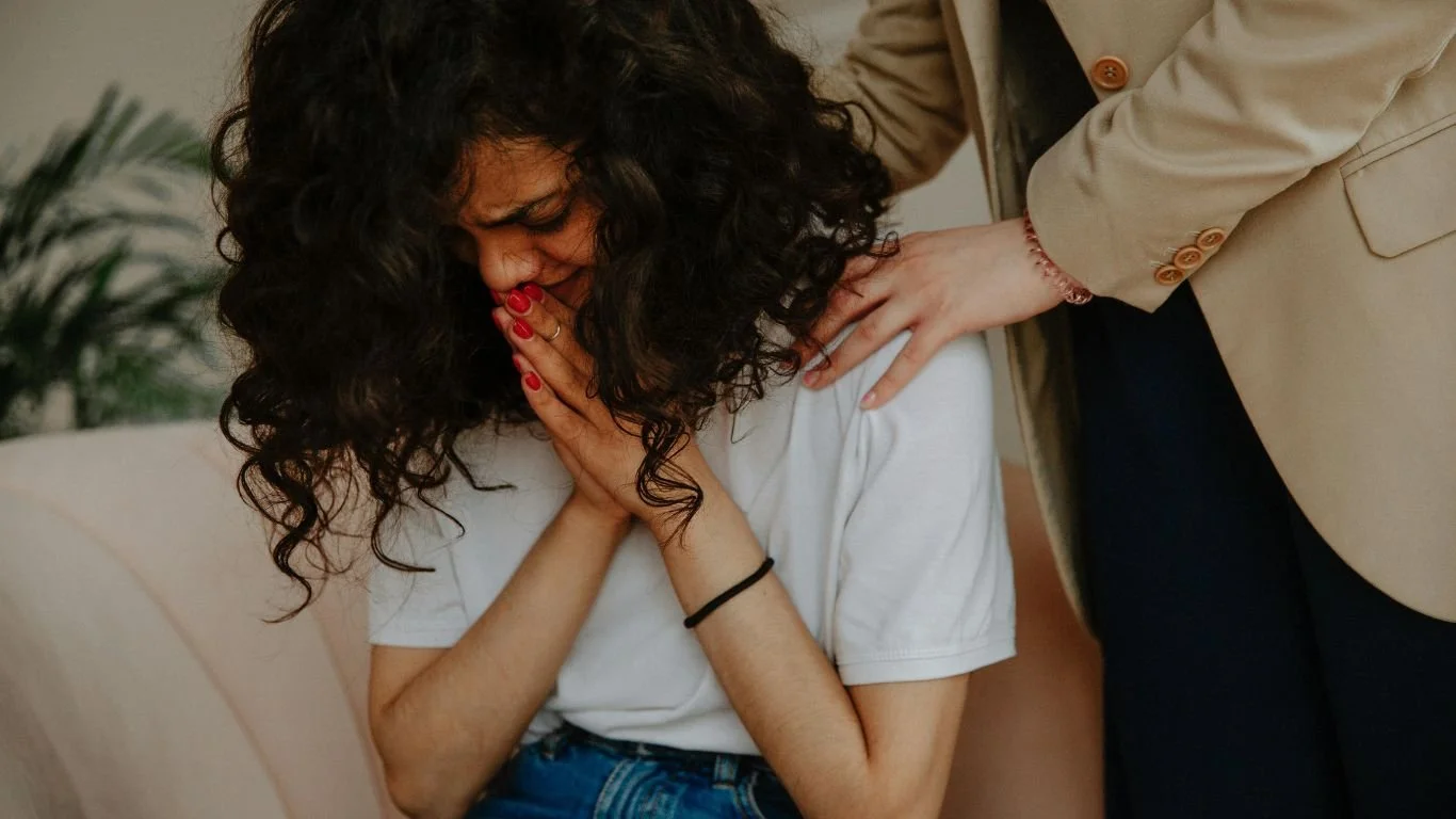 A woman with curly hair is holding her face in her hands, appearing distressed, while another woman in a beige blazer comforts her by placing a hand on her shoulder.