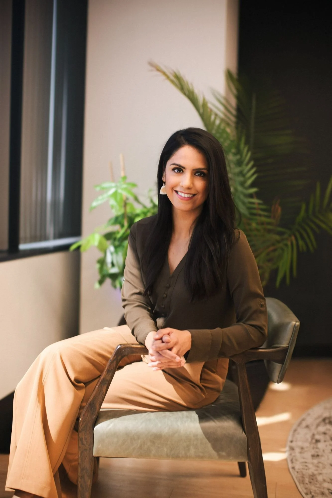 A woman sitting on a chair indoors with a smile, dark hair, wearing a dark blouse and tan pants, with green plants in the background.
