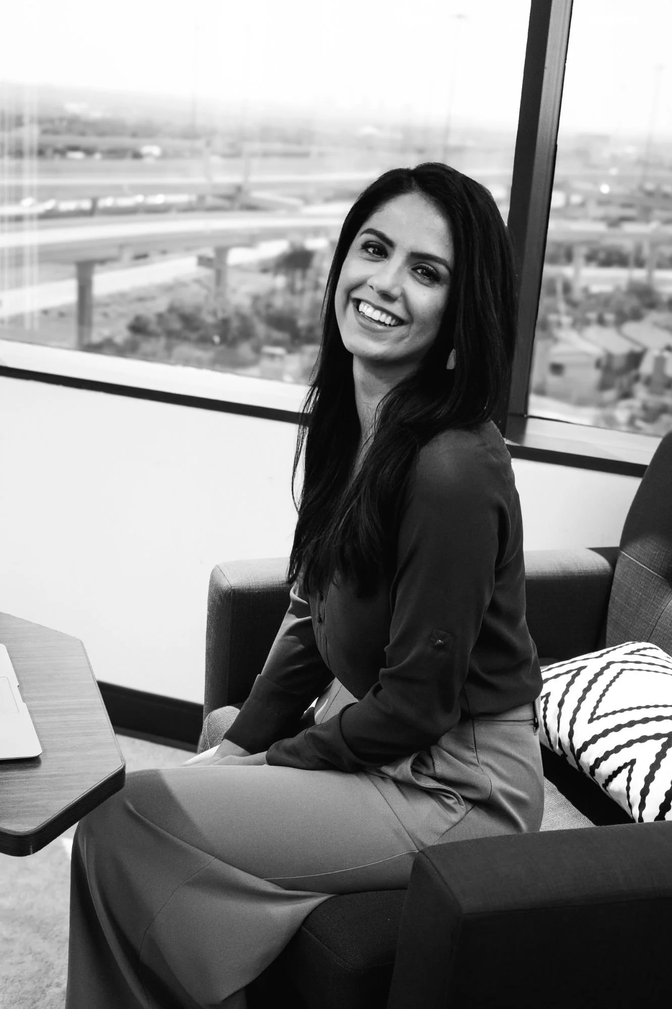 A smiling woman with long dark hair sitting in an office chair next to a window overlooking a highway and cityscape, dressed in professional attire.
