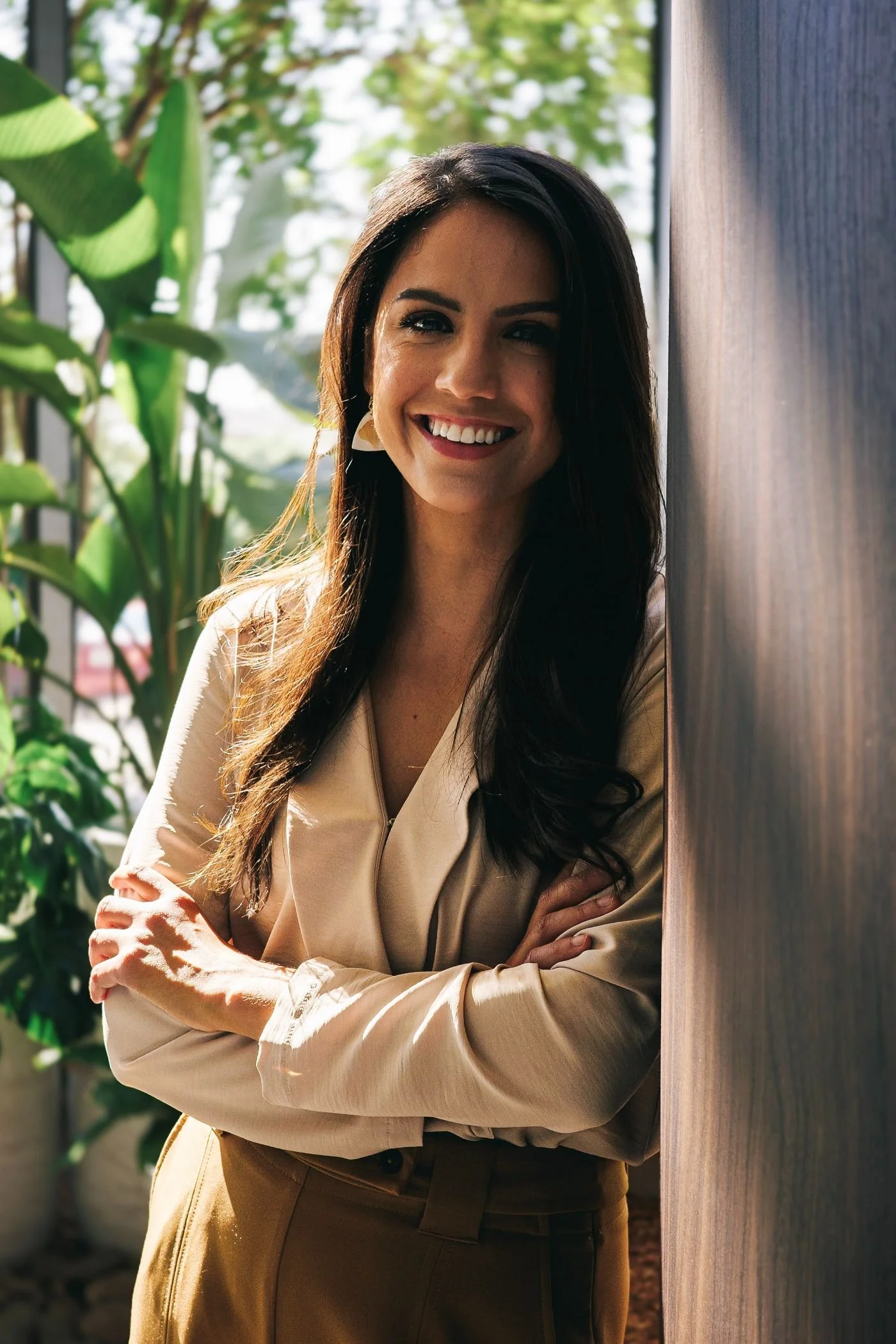 A woman with long dark hair and a beige blouse, smiling and standing next to a wooden wall outdoors with green plants in the background.